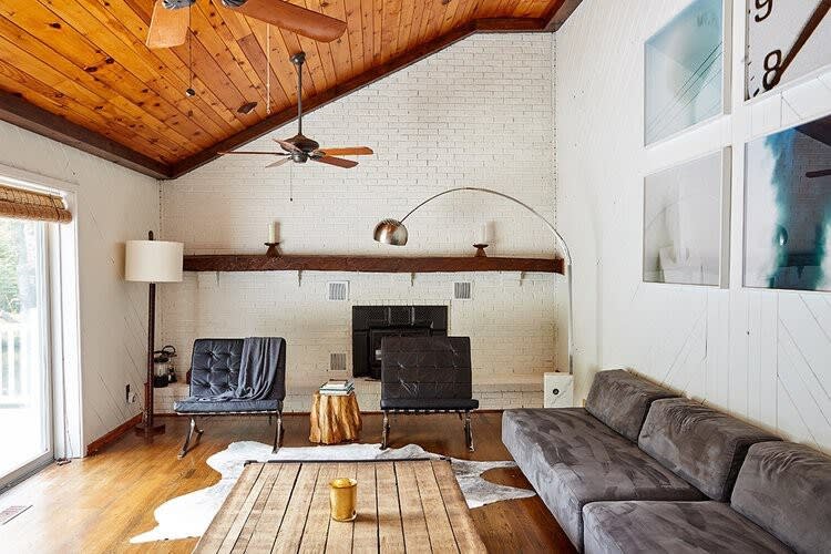 Spacious living room with a vaulted wood-paneled ceiling and a white brick fireplace. Dark leather chairs with faux fur throws face a large wooden coffee table, with an arched floor lamp providing light.