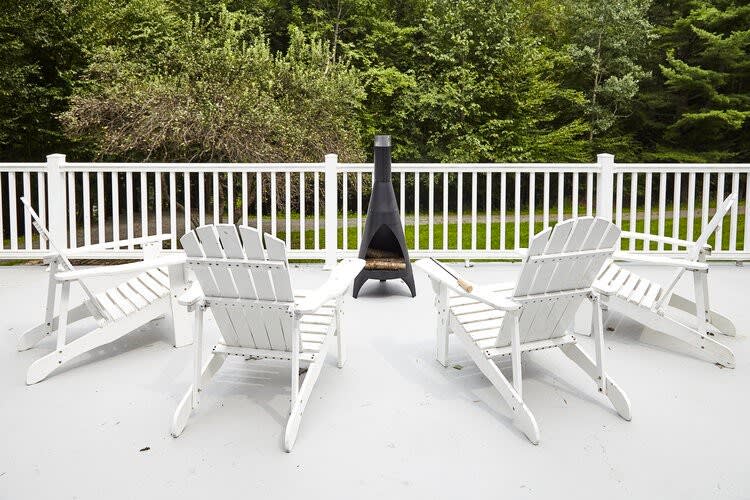 Outdoor fire pit area on a grey deck, surrounded by four white Adirondack chairs. A modern black chiminea serves as the fire pit, set against a backdrop of lush green trees and a white railing.