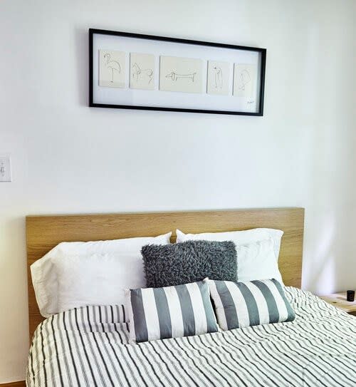 A close-up view of a bed in a bright bedroom, featuring a light wooden headboard, striped duvet, and a combination of gray textured and striped throw pillows. A framed art piece with minimalist drawings hangs on the white wall above.