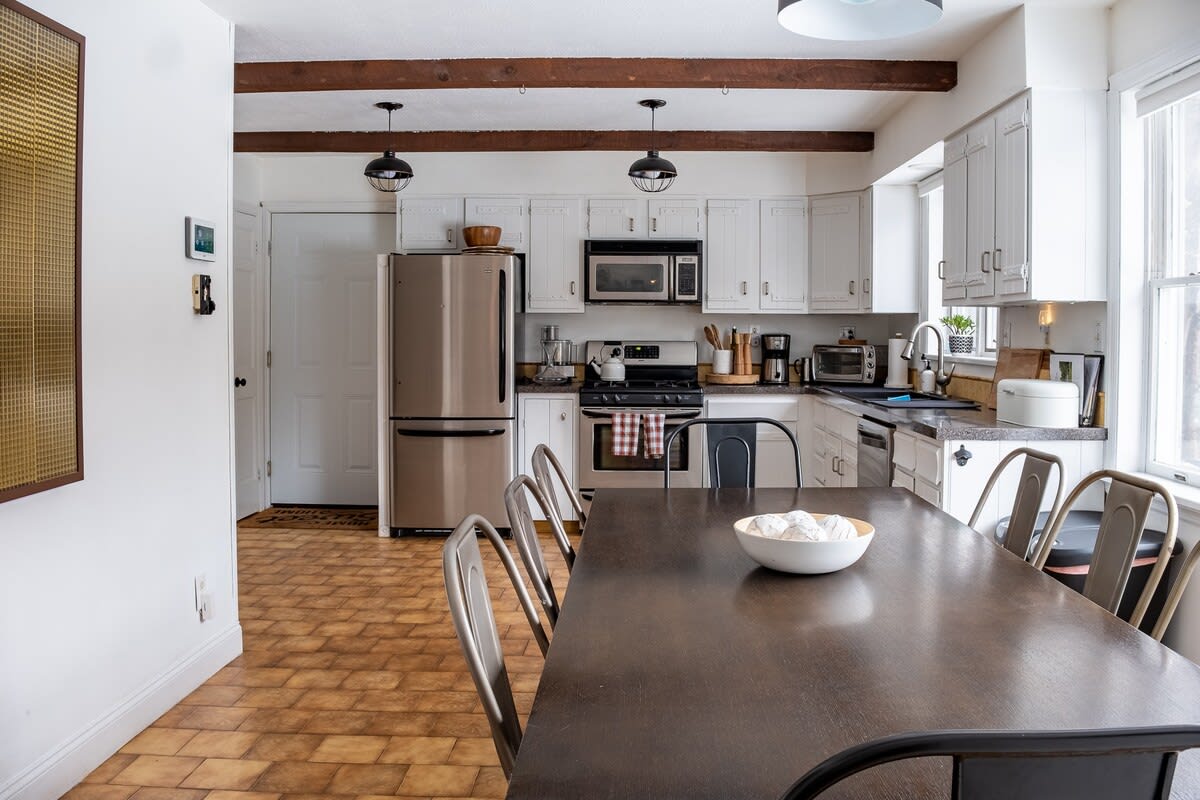 A rustic-modern dining area with a large dark wooden table and metal chairs, leading into a white kitchen with stainless steel appliances, exposed wooden beams, and patterned tile flooring.