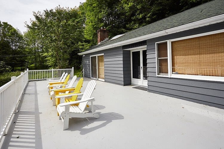 Spacious grey deck with a white railing, featuring a row of yellow and white Adirondack chairs. The deck overlooks a wooded area, providing a serene outdoor lounging space.