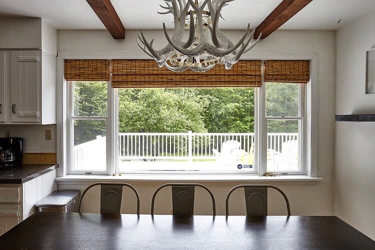 A dining area with a dark rectangular table and metal chairs, positioned in front of a wide window with bamboo blinds, offering a view of a green outdoor deck and trees. A unique antler-style chandelier hangs above.
