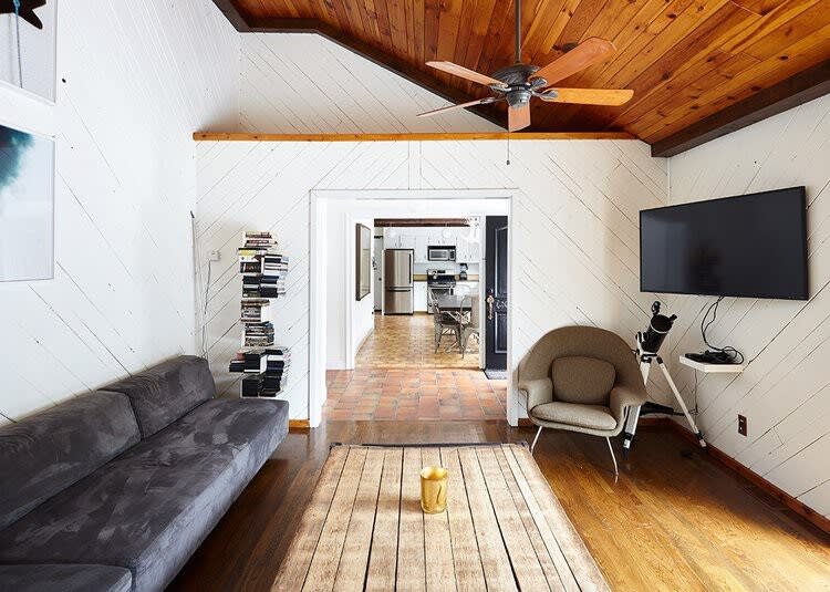 Living room with a dark grey sectional sofa, a large wooden coffee table, and a wall-mounted TV. The room features white shiplap walls, a wood-paneled vaulted ceiling, and an opening leading to the kitchen/dining area.