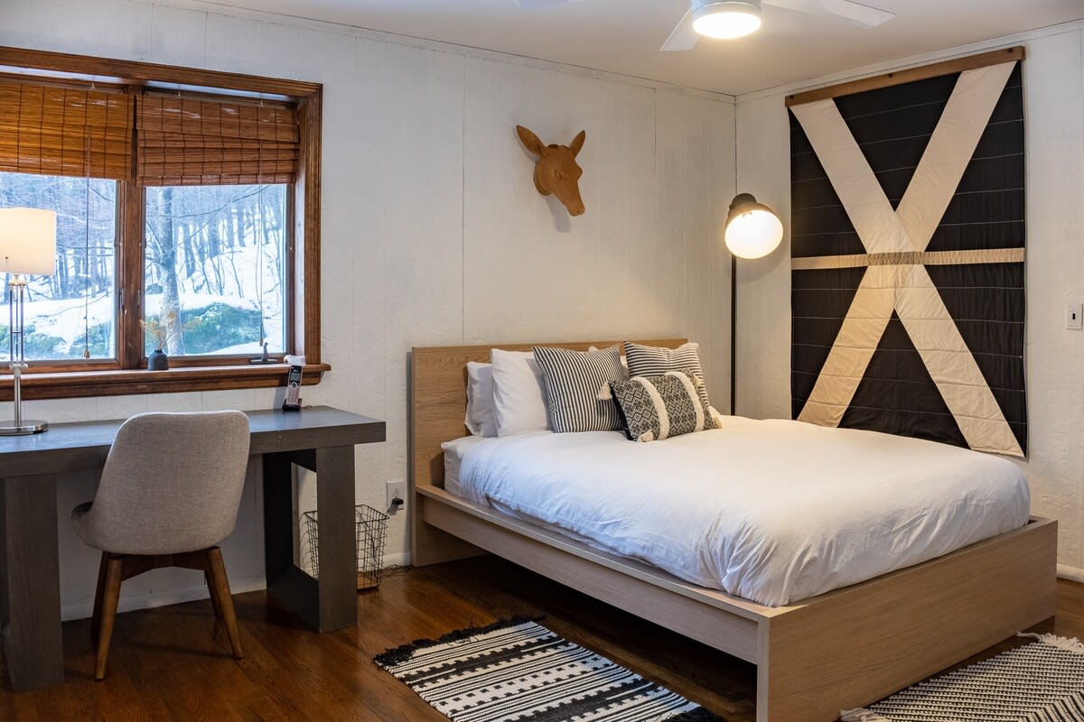 A cozy bedroom with a light wood platform bed, white bedding, and patterned throw pillows, featuring a desk and chair by a window with bamboo blinds overlooking a snowy landscape. A large black and white geometric tapestry adorns the wall.