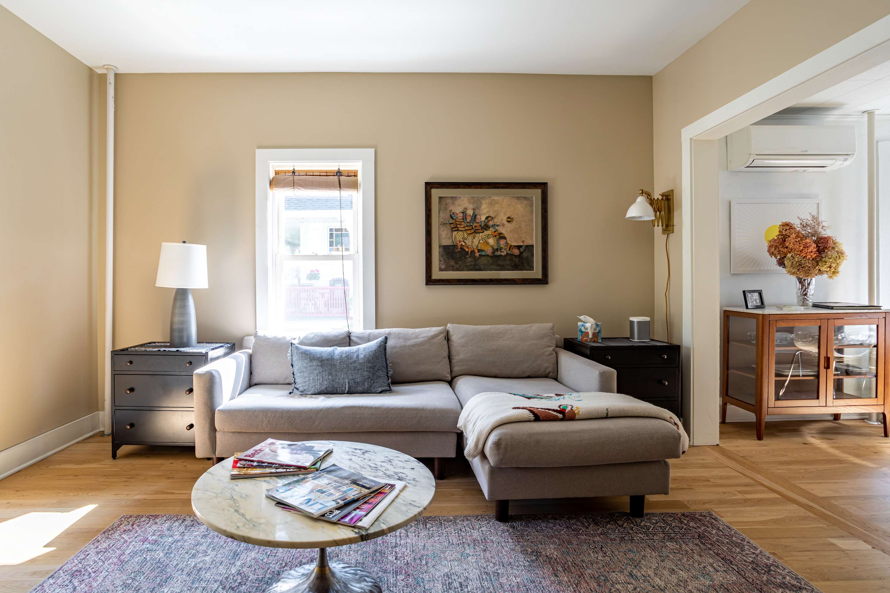 A cozy living room with light beige walls, a light gray sectional sofa with a chaise lounge, a round marble coffee table, and a patterned area rug. A window with wooden blinds is visible on the left, and a framed painting hangs above the sofa.