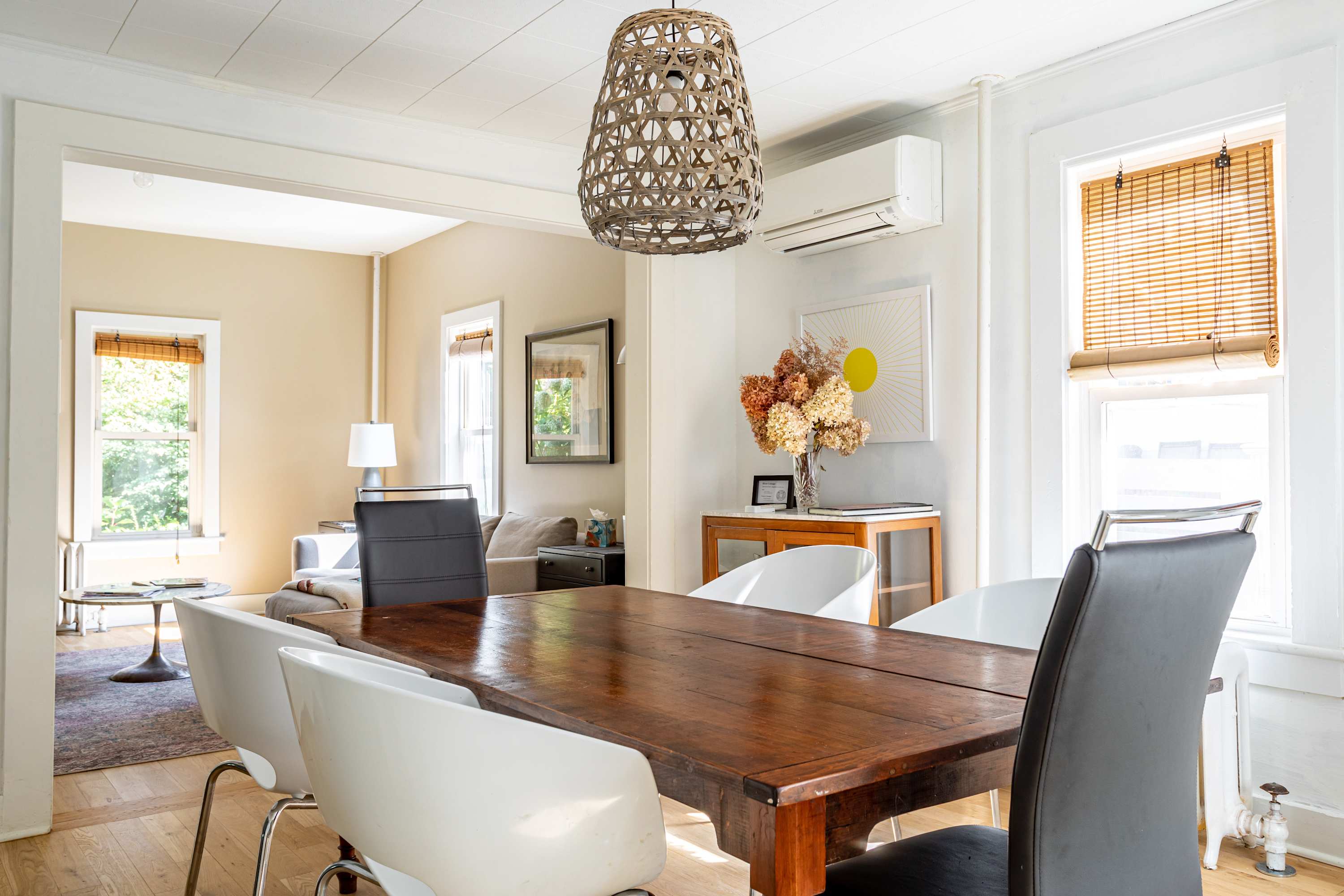 A view from the dining room looking into the living room, showing a rustic wooden dining table with white and black chairs, and a woven pendant light. The living room features a light gray sectional sofa and a marble coffee table.