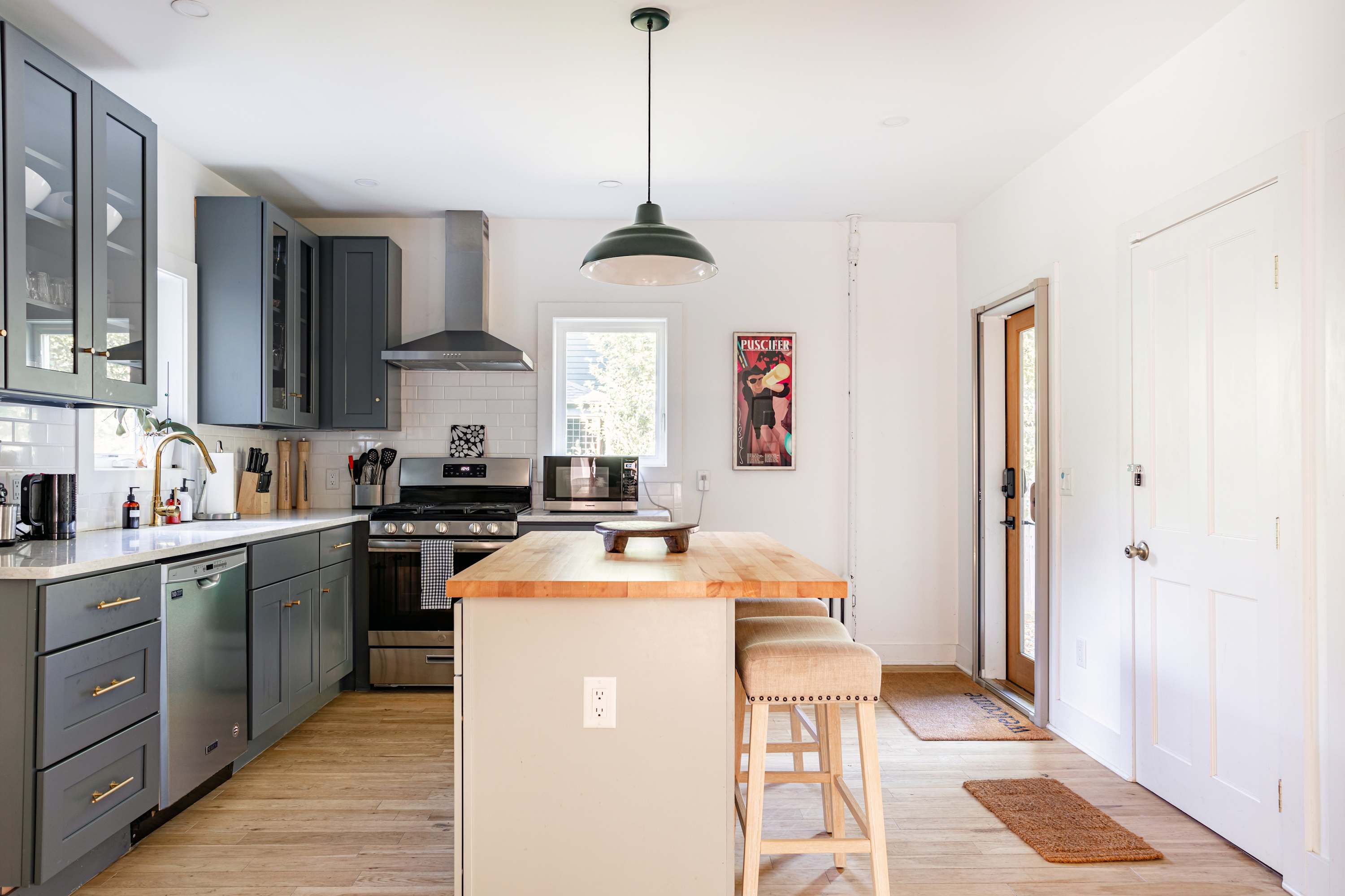 A full view of the kitchen showing gray shaker-style cabinets, white subway tile backsplash, and stainless steel appliances including a range and range hood. A kitchen island with a butcher block top and two bar stools is in the foreground.