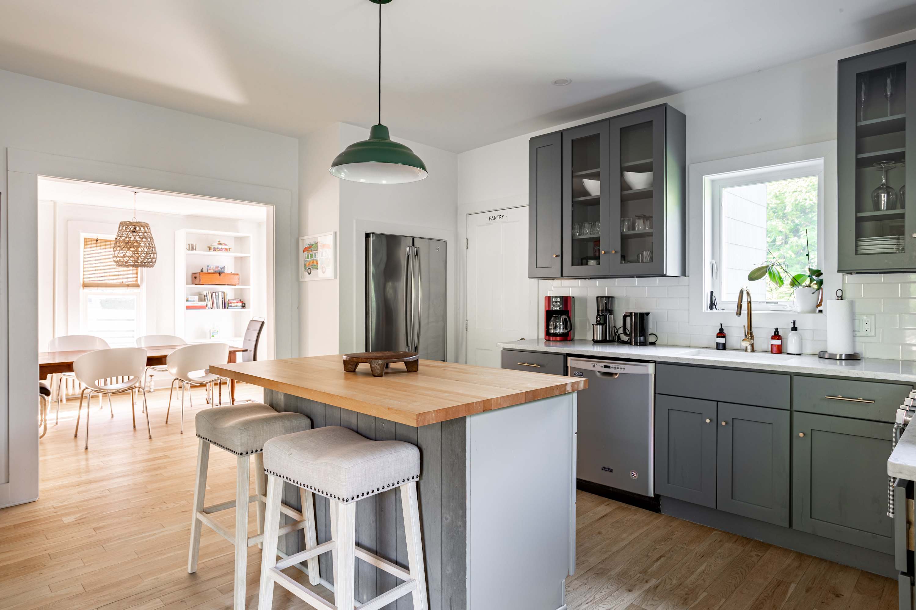 A bright kitchen with white walls, gray shaker-style cabinets, white subway tile backsplash, and light wood floors. A kitchen island with a butcher block countertop and two bar stools is in the foreground, and a stainless steel refrigerator is visible.