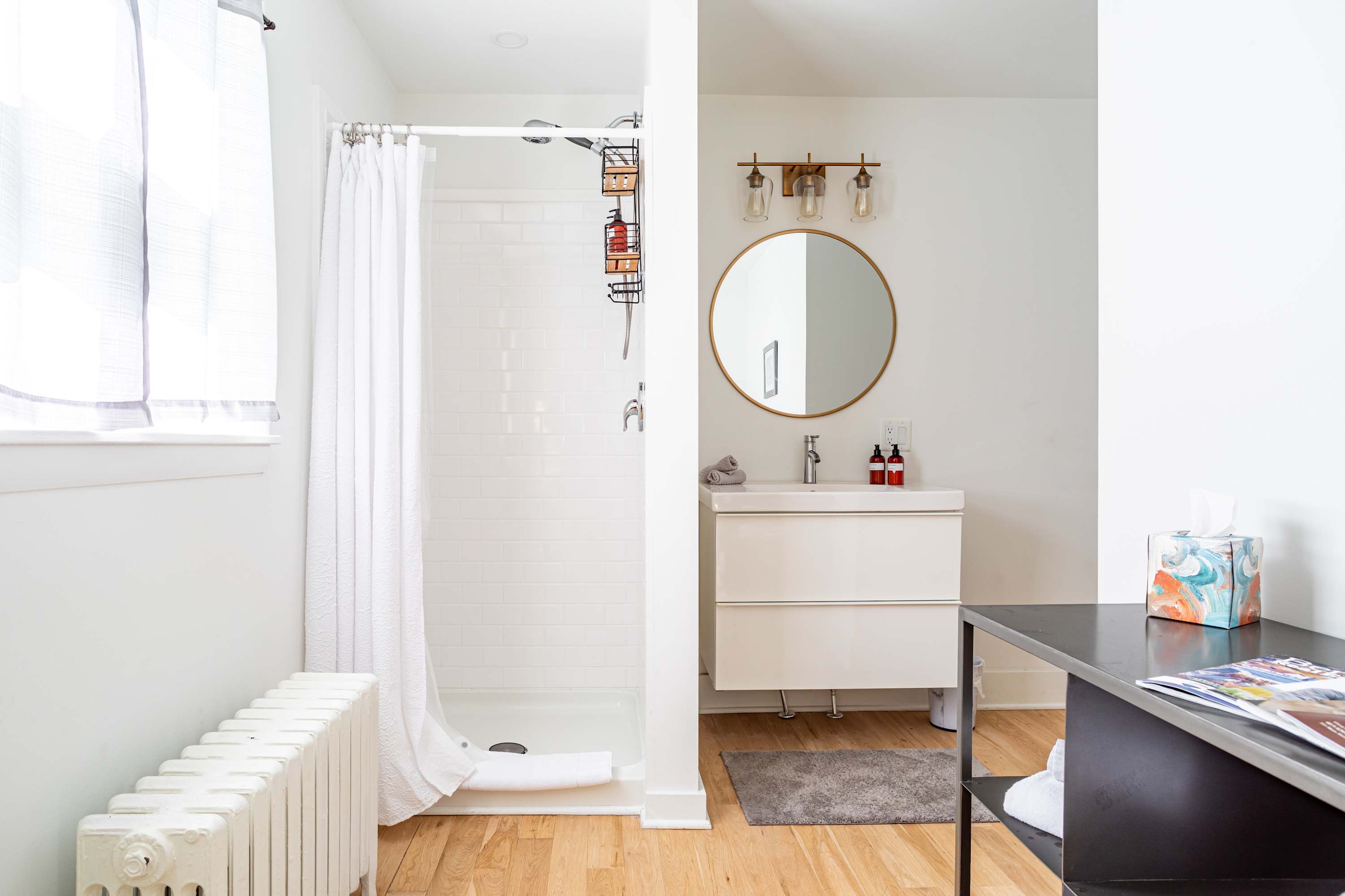 A closer view of the bathroom showing the walk-in shower with a white curtain and subway tiles, next to a window. A modern vanity with a round mirror and brass light fixture is visible, along with a white radiator and a black metal shelving unit.