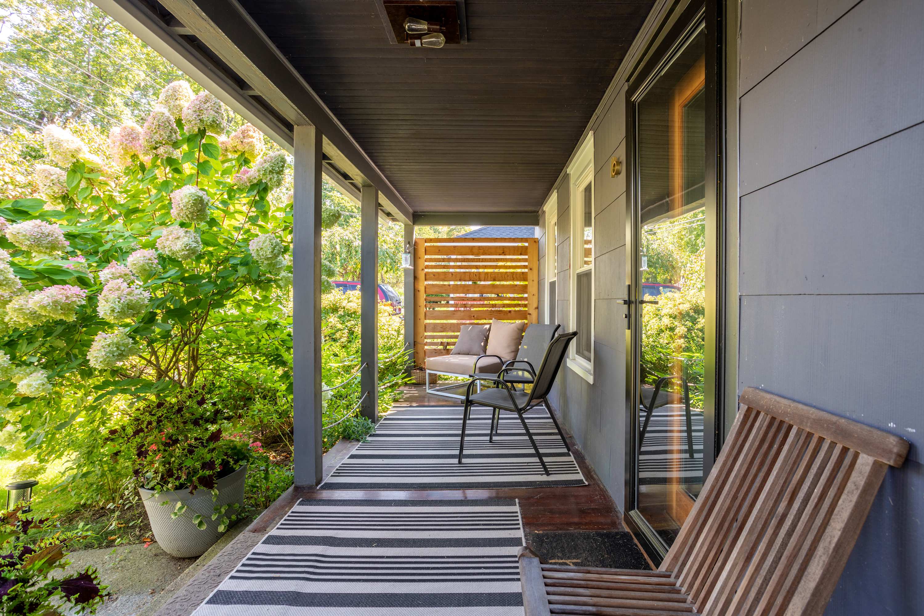 A long, covered front porch with dark gray siding, a black and white striped rug, and various seating options including a wooden chair and a small sofa, surrounded by lush greenery.