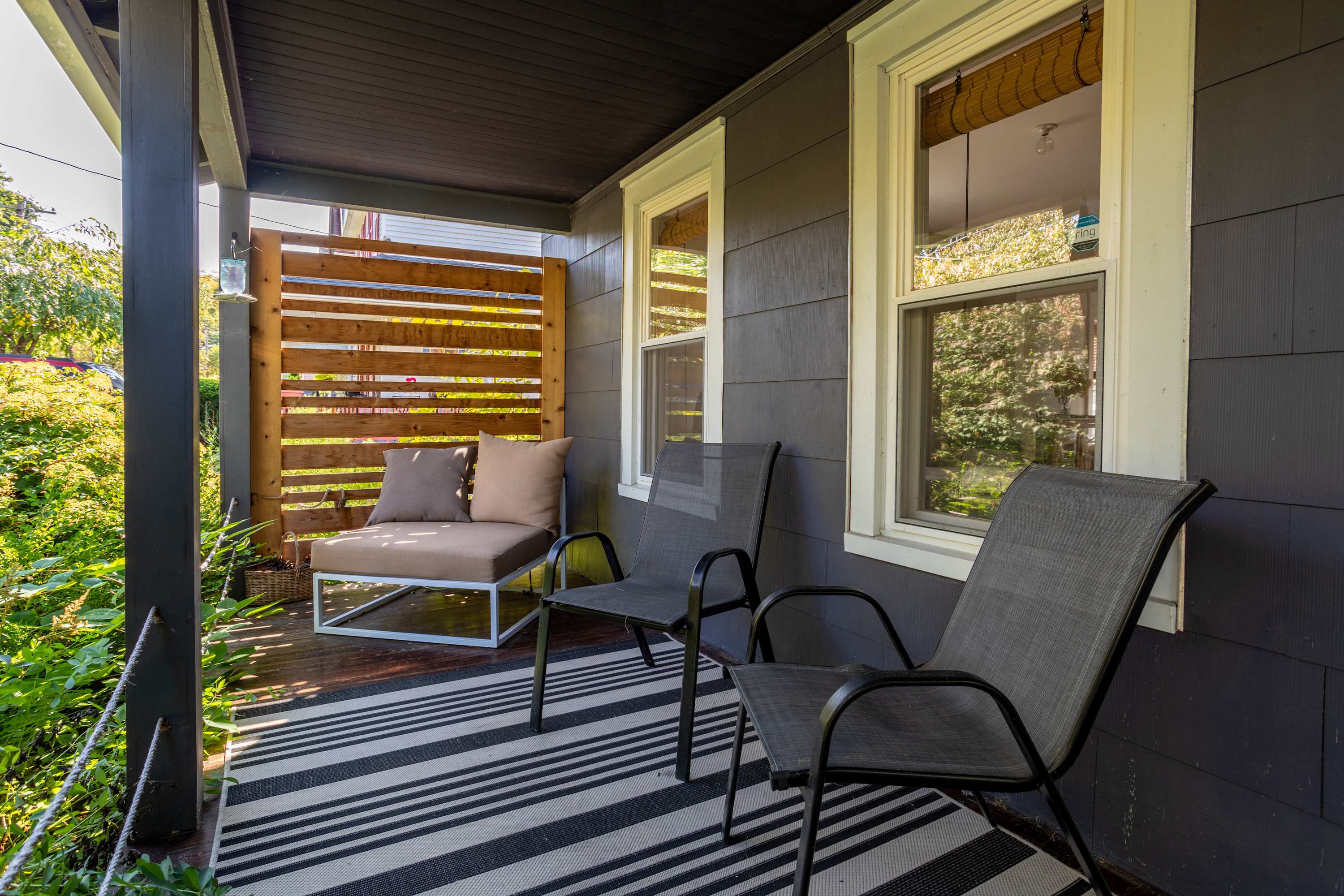A cozy covered porch features dark gray siding and two large windows, furnished with a striped outdoor rug, two dark metal chairs, and a light brown outdoor sofa with cushions. A slatted wooden privacy screen adds a touch of warmth and seclusion.