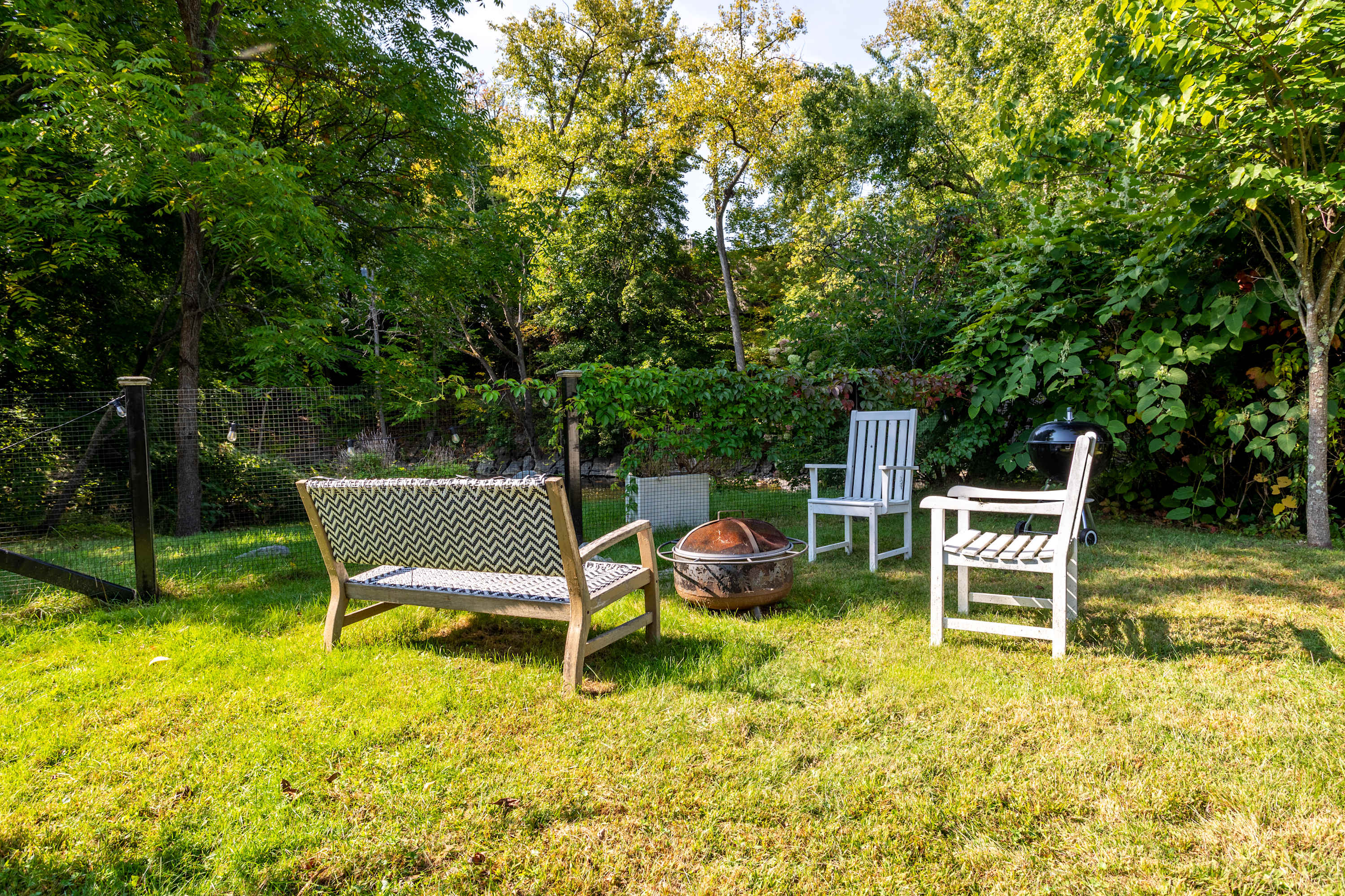 A grassy backyard area set up for relaxation and entertaining, featuring a wooden bench, a white chair, a grill, and a fire pit, all surrounded by dense green trees and fencing.