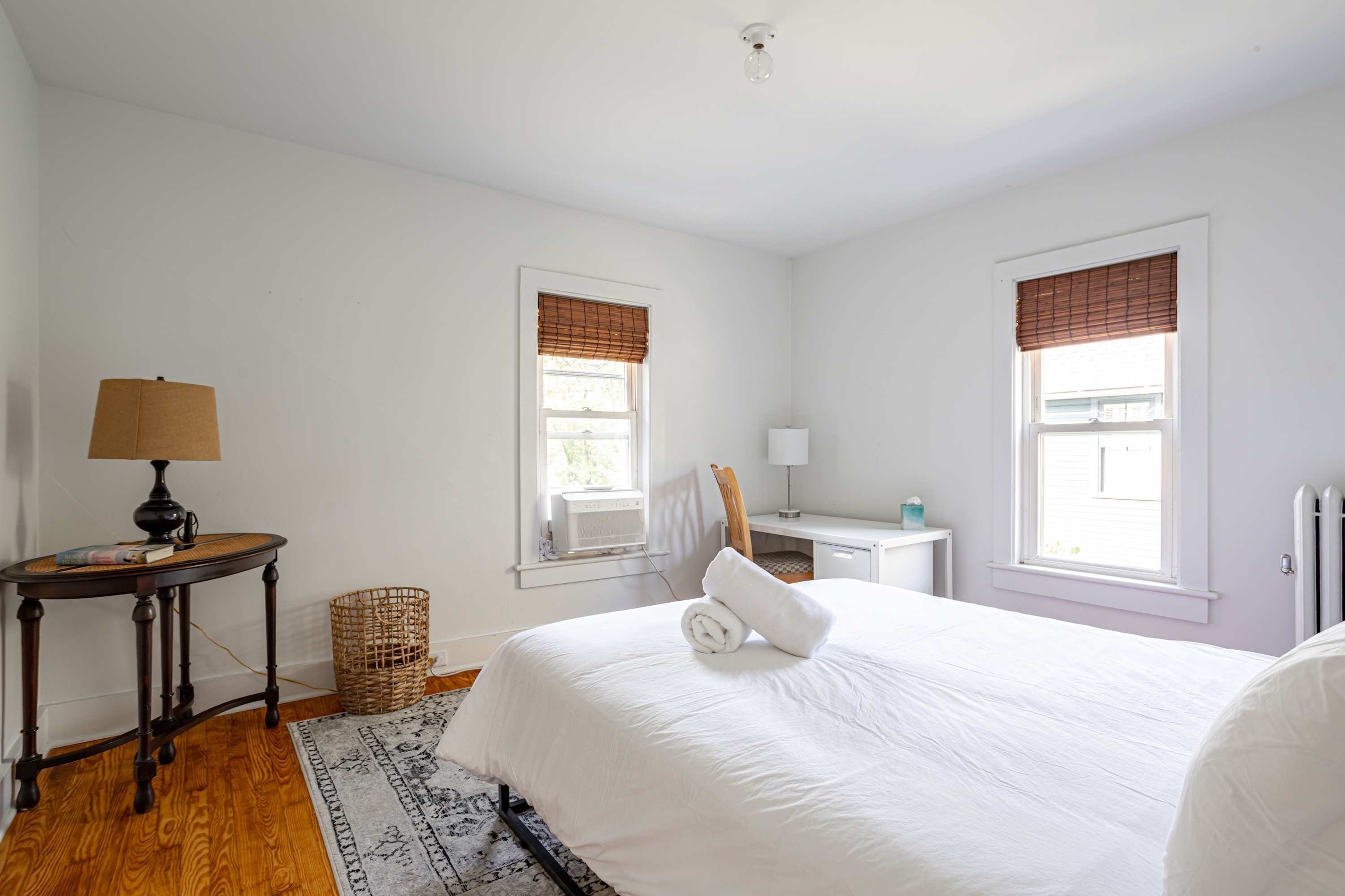 Another view of the bright bedroom shows the queen bed with white linens and rolled towels, alongside a round wooden side table with a lamp. Two windows with bamboo blinds provide natural light.