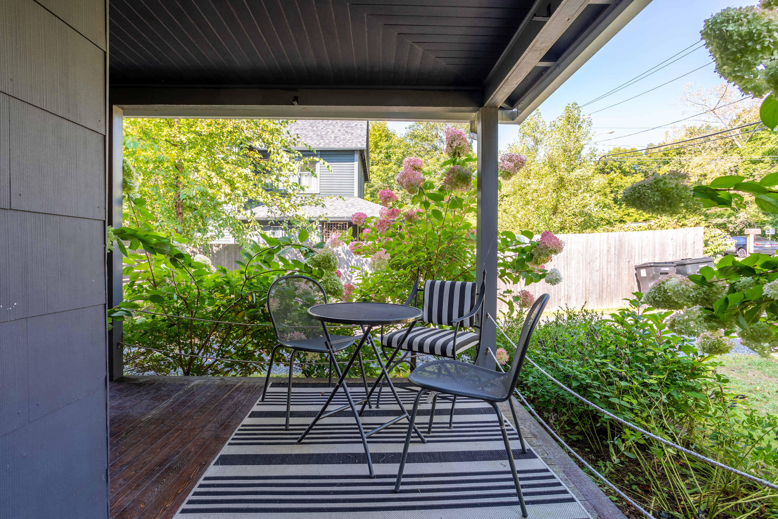 This inviting covered porch offers a small black metal bistro table with two matching chairs, one featuring a striped cushion, all resting on a black and white striped outdoor rug. Lush green bushes and vibrant pink hydrangeas frame the outdoor seating area.