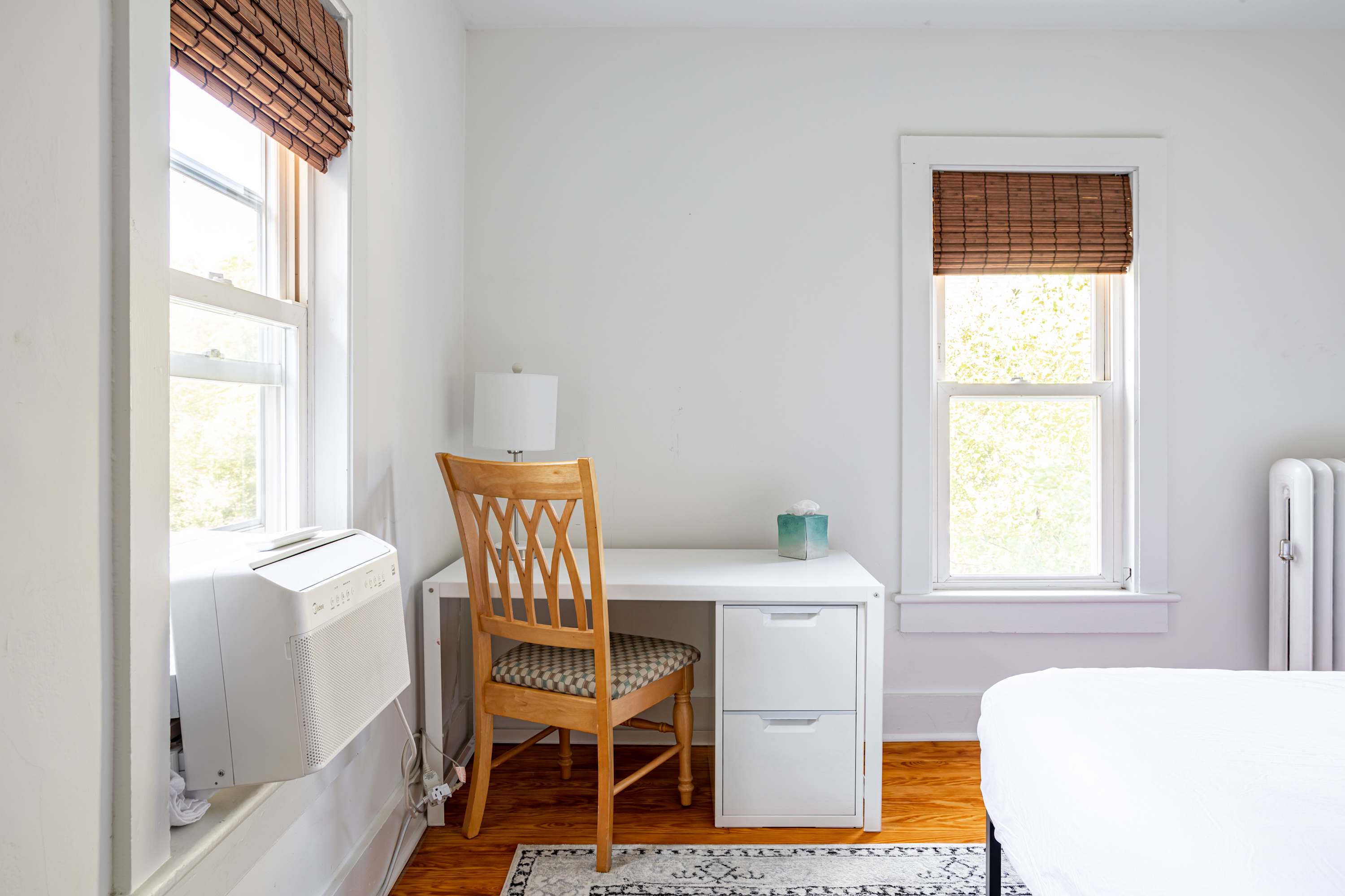 A dedicated workspace is visible in the bright bedroom, featuring a white desk with drawers and a wooden chair. Two windows with bamboo blinds frame the desk, and a small white mini-fridge is tucked underneath.