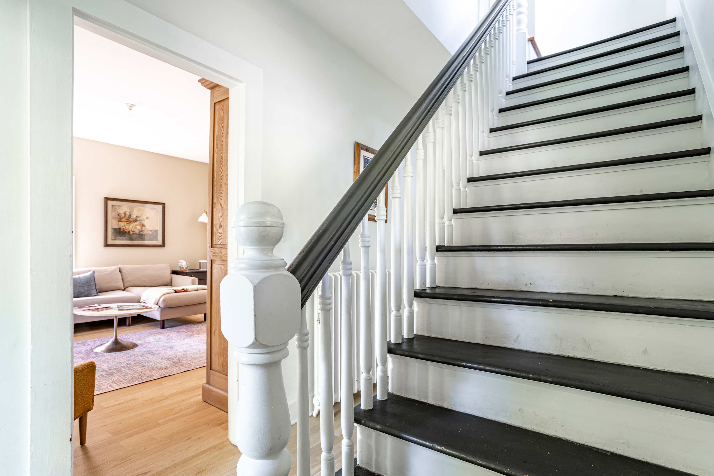 A bright foyer features a white staircase with dark treads and a dark handrail, leading upstairs. Through an open doorway, a cozy living room with a light-colored sofa and a round coffee table is visible.