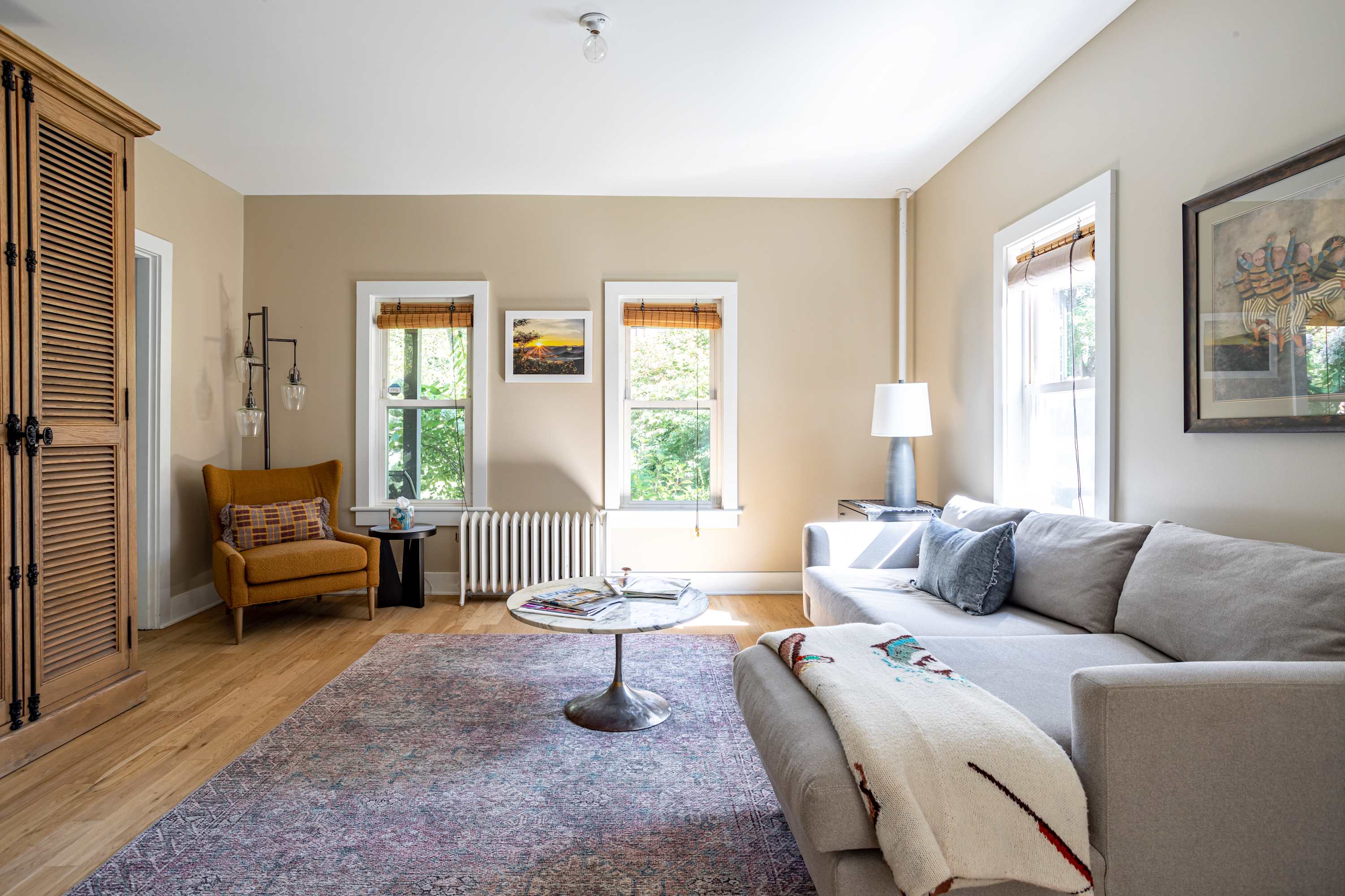 A wider view of the living room showing a light gray sectional sofa, a round marble coffee table, and a patterned area rug. To the left, there's a mustard yellow armchair, a tall wooden cabinet, and two windows with wooden blinds.