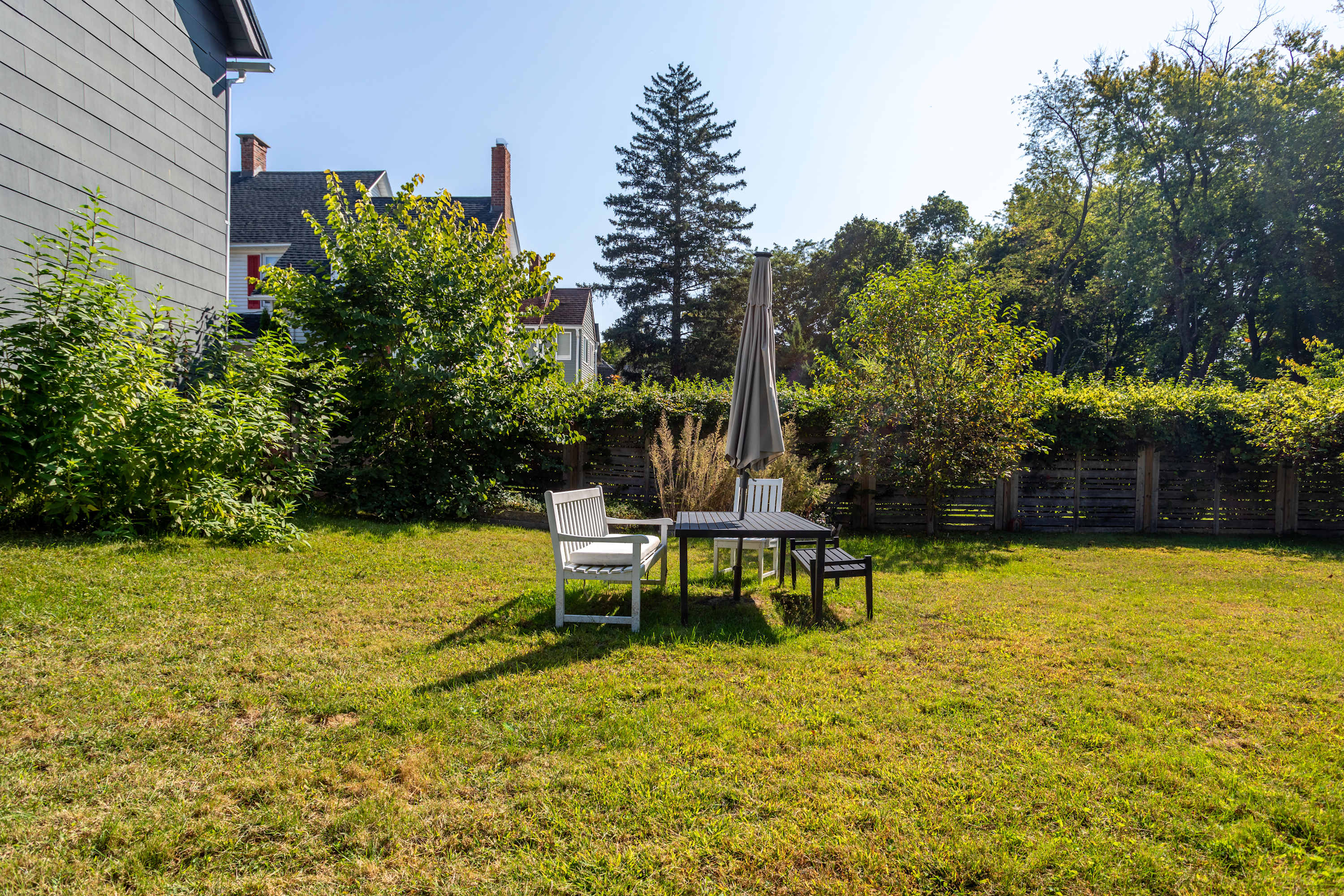 A well-maintained grassy backyard features a black outdoor dining table with two white chairs and a black bench, shaded by a large gray umbrella. Mature trees and a wooden fence define the perimeter of this sunny outdoor space.