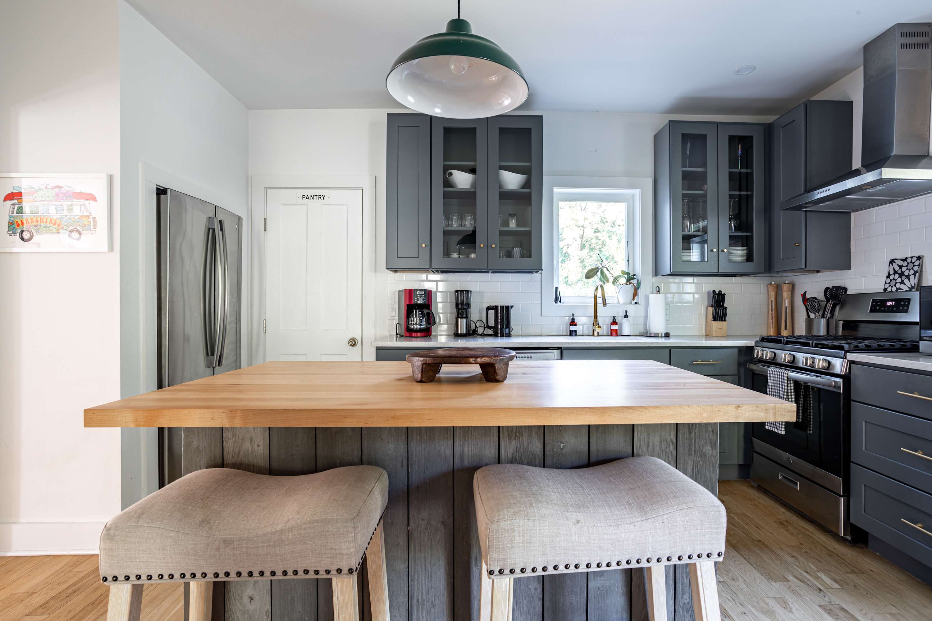 A wide shot of the kitchen featuring gray shaker-style cabinets, white countertops, and a white subway tile backsplash. A kitchen island with a butcher block top and two bar stools is central, with a stainless steel refrigerator and a "Pantry" door visible.