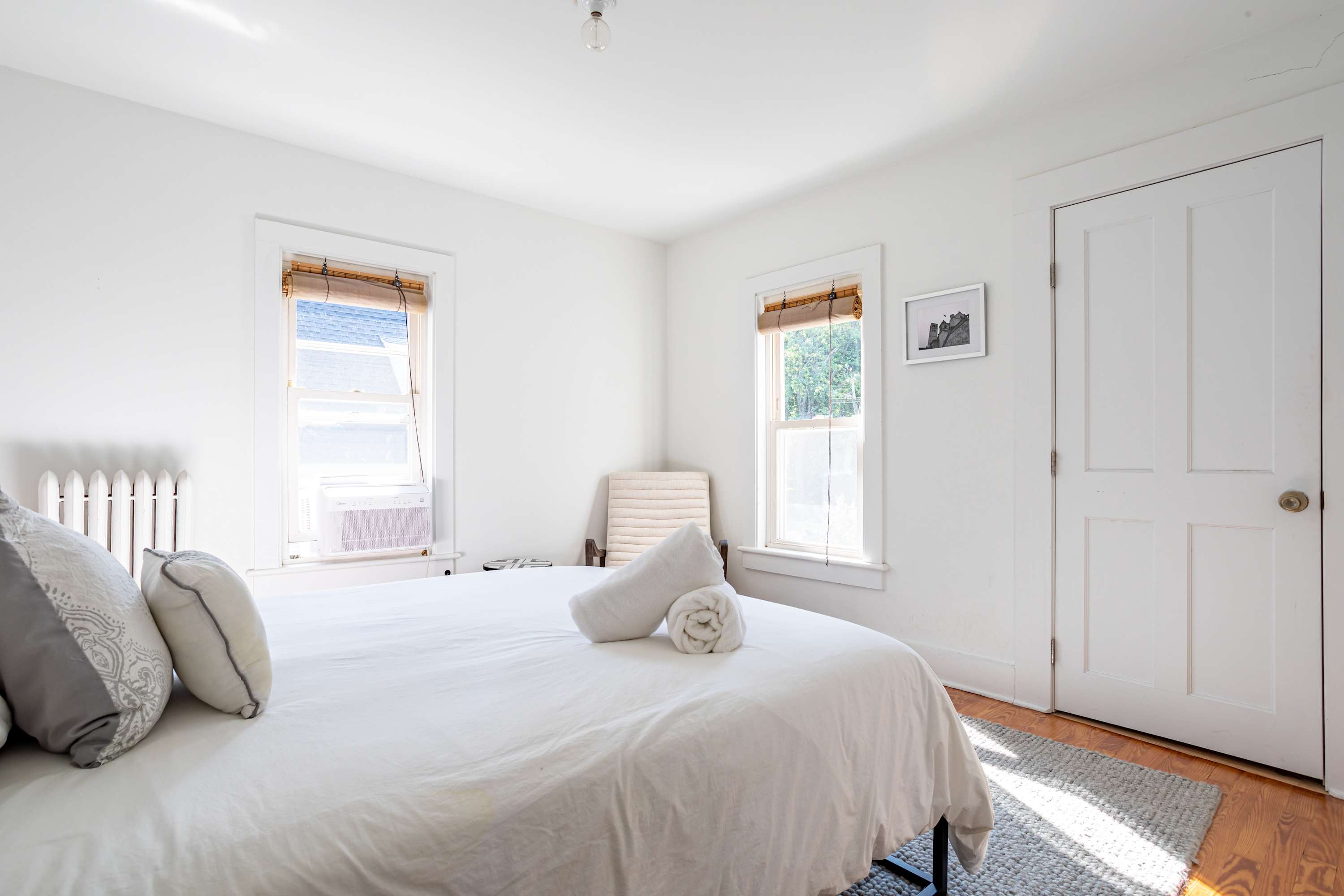 This bright bedroom showcases a queen bed with white linens and rolled towels, positioned on a light grey rug. Two windows with natural bamboo blinds allow ample light, and a light-colored armchair is visible in the corner.