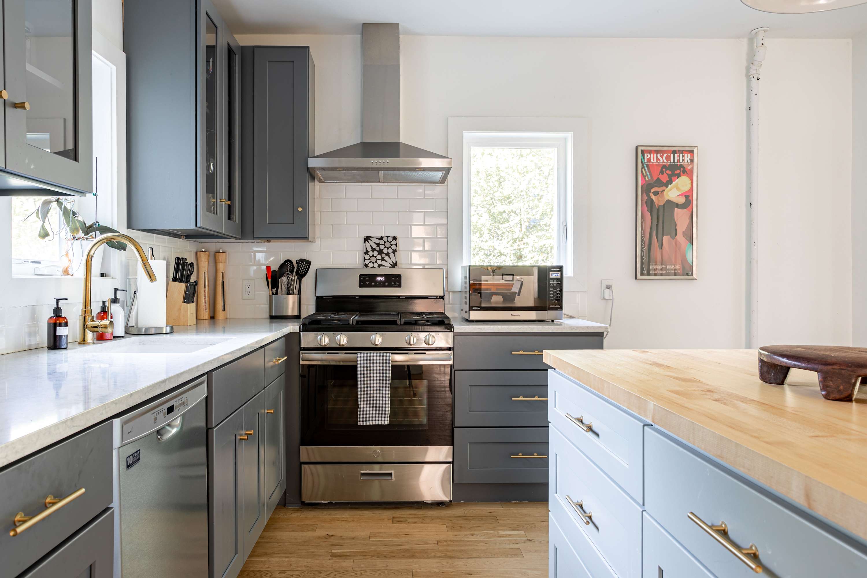 A close-up view of the kitchen showing the stainless steel range, range hood, and microwave, flanked by gray cabinets and white subway tile backsplash. The kitchen island with its butcher block top is partially visible on the right.