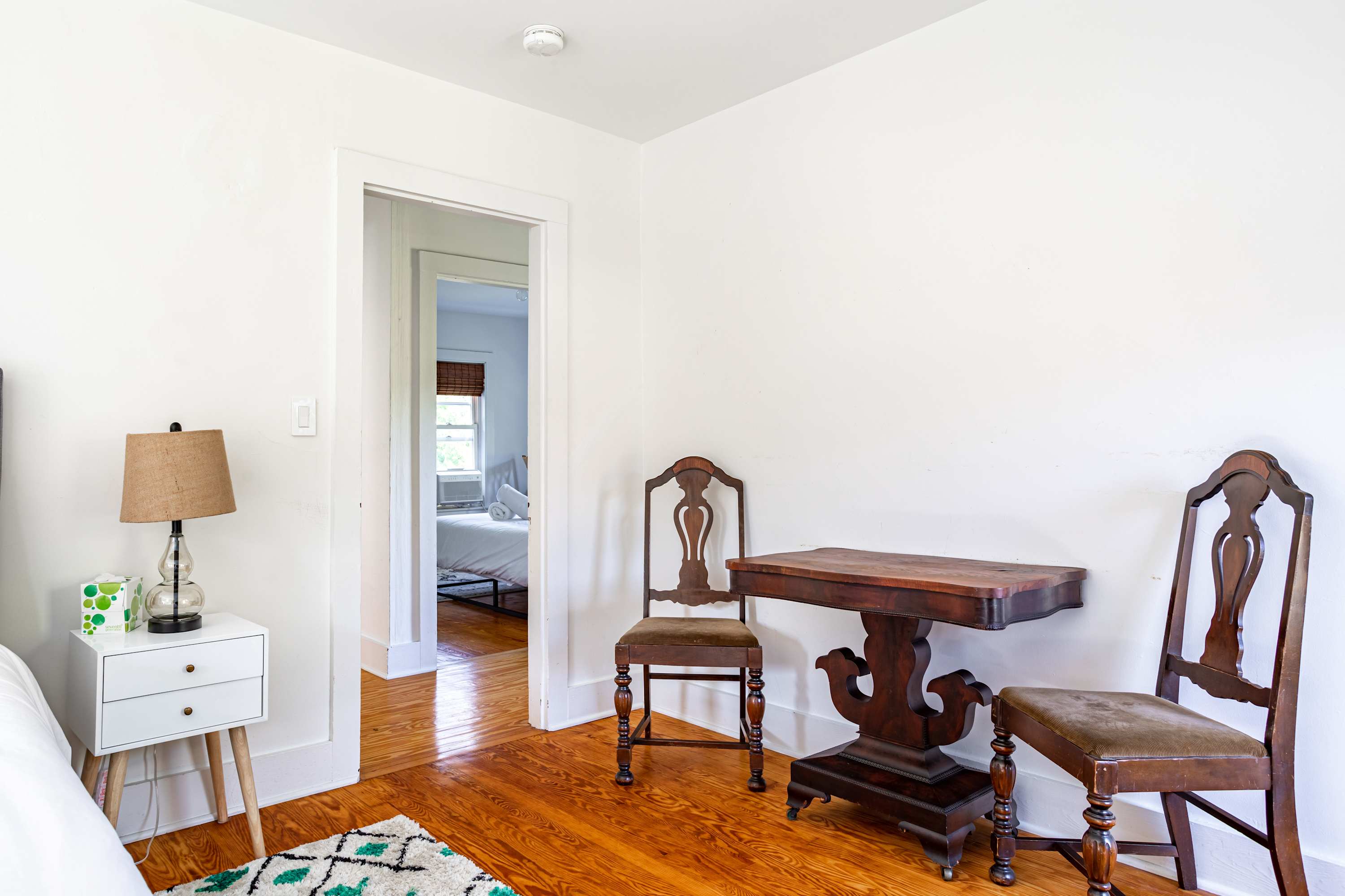 A small transitional area with polished hardwood floors features a dark wooden console table and two antique-style chairs. An open doorway in the background leads into another bright bedroom.