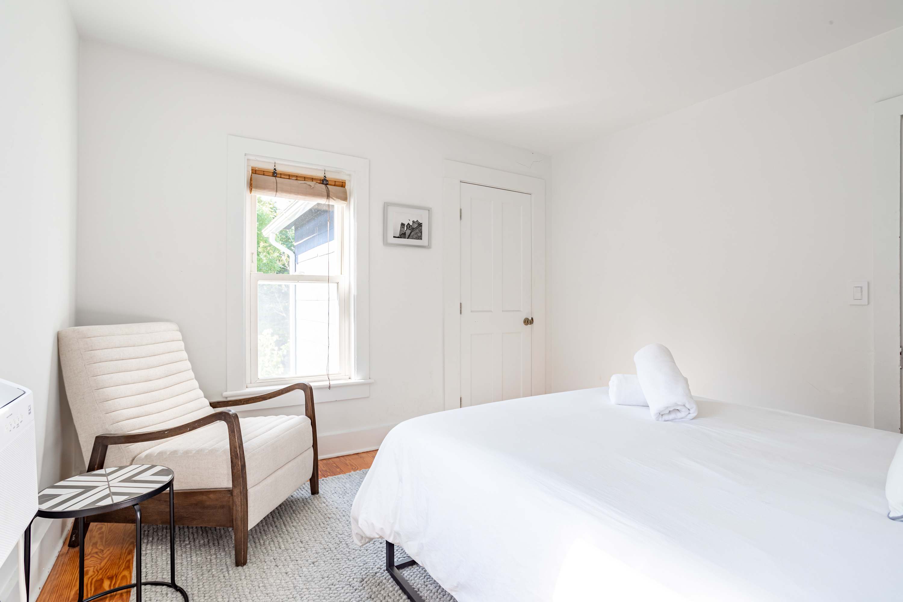 A corner view of the bright bedroom highlights a light-colored armchair with wooden arms next to a small round side table with a geometric pattern. The queen bed with white linens and rolled towels is partially visible on a grey rug.