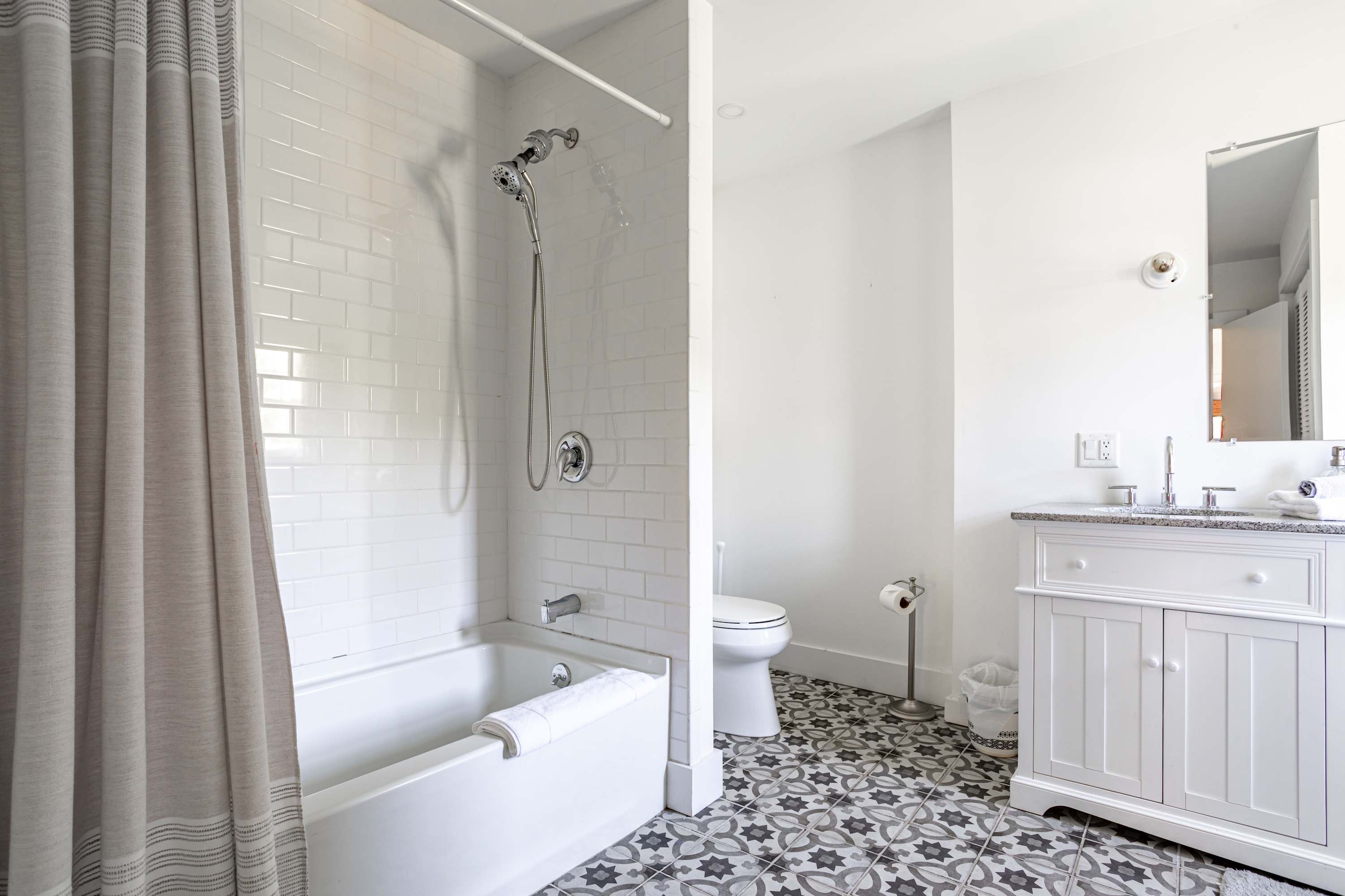 A modern bathroom features a white subway-tiled shower/tub combo with a grey shower curtain. The floor is adorned with patterned grey and white tiles, and a white vanity with a sink and mirror is visible on the right.