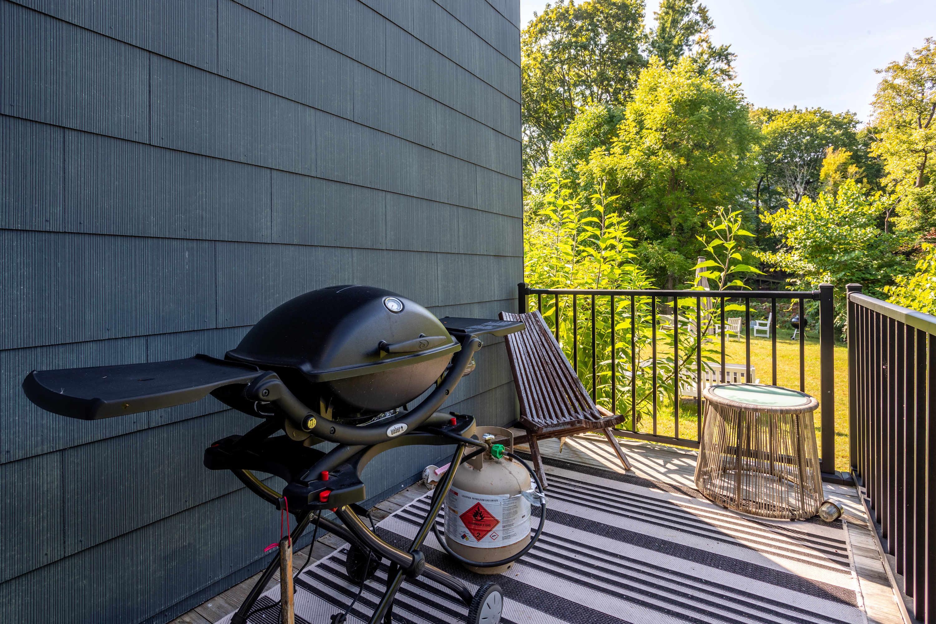 A compact outdoor deck, adjacent to dark gray siding, features a black Weber BBQ grill with a propane tank, a wooden slatted lounge chair, and a small round side table. A black and white striped rug adds a decorative touch to the grilling area.