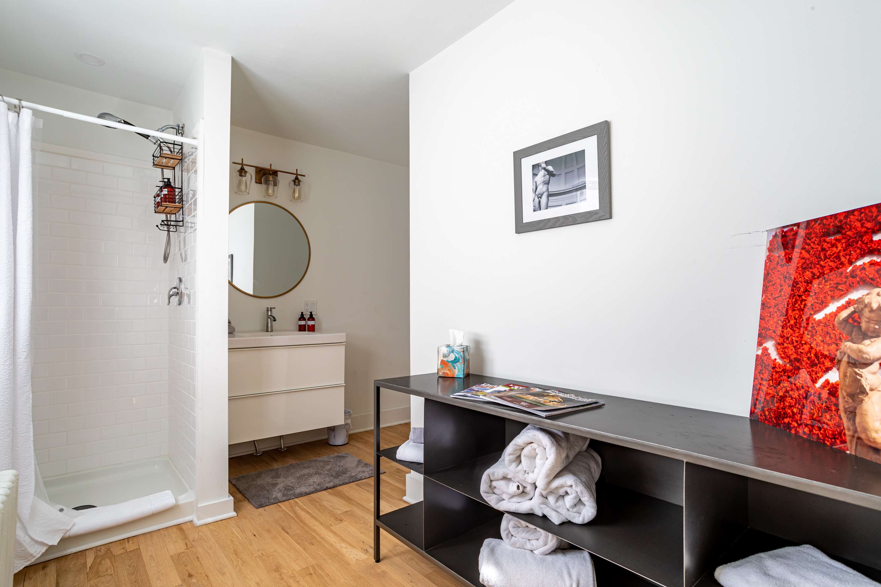 A bright bathroom with white walls and light wood floors, featuring a walk-in shower with white subway tiles and a glass partition. A modern vanity with a round mirror and brass light fixture is visible, along with a black metal shelving unit.