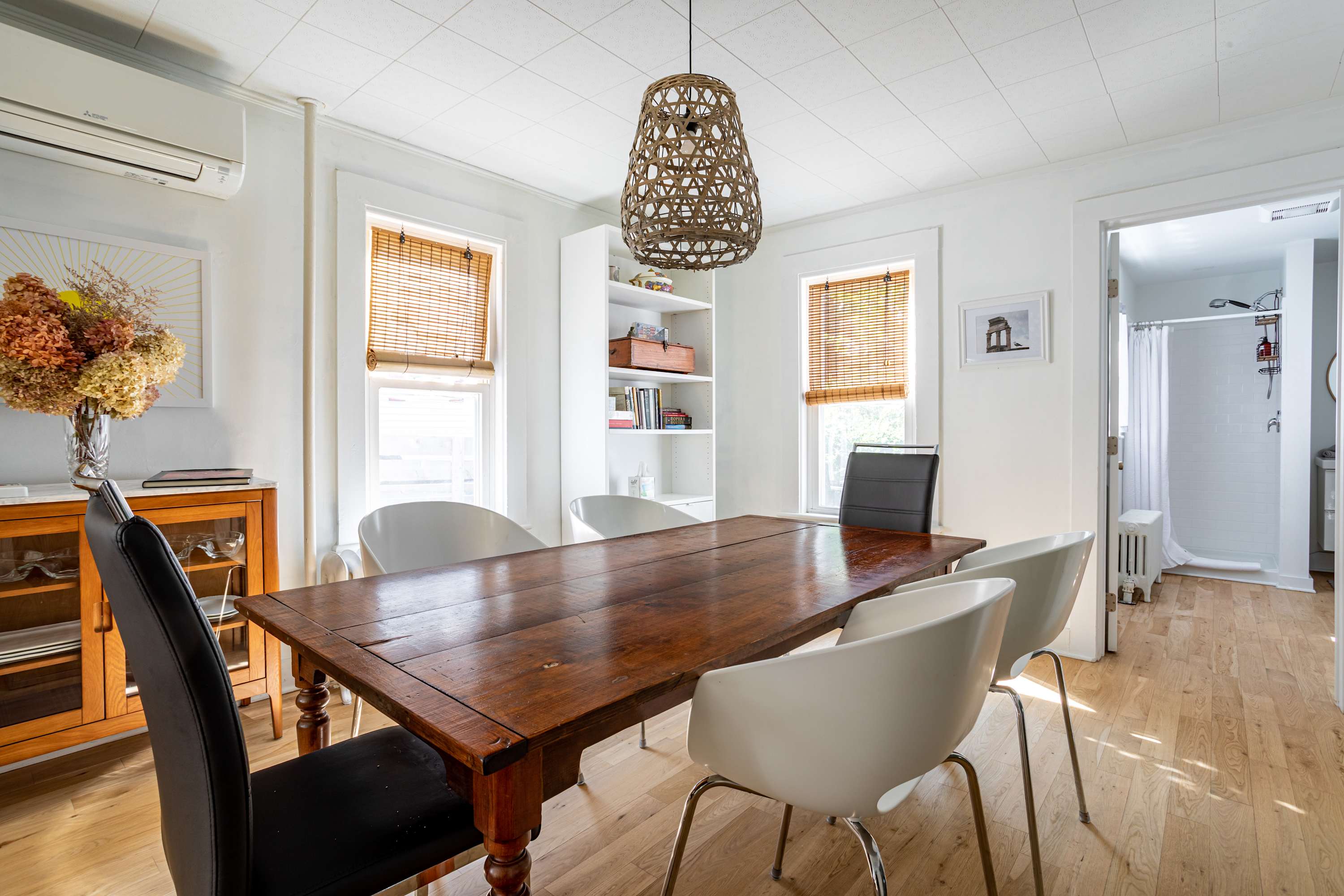 A dining room with white walls, a rustic wooden dining table, and a mix of white and black dining chairs. A woven pendant light hangs above the table, and a built-in bookshelf is visible on the left.