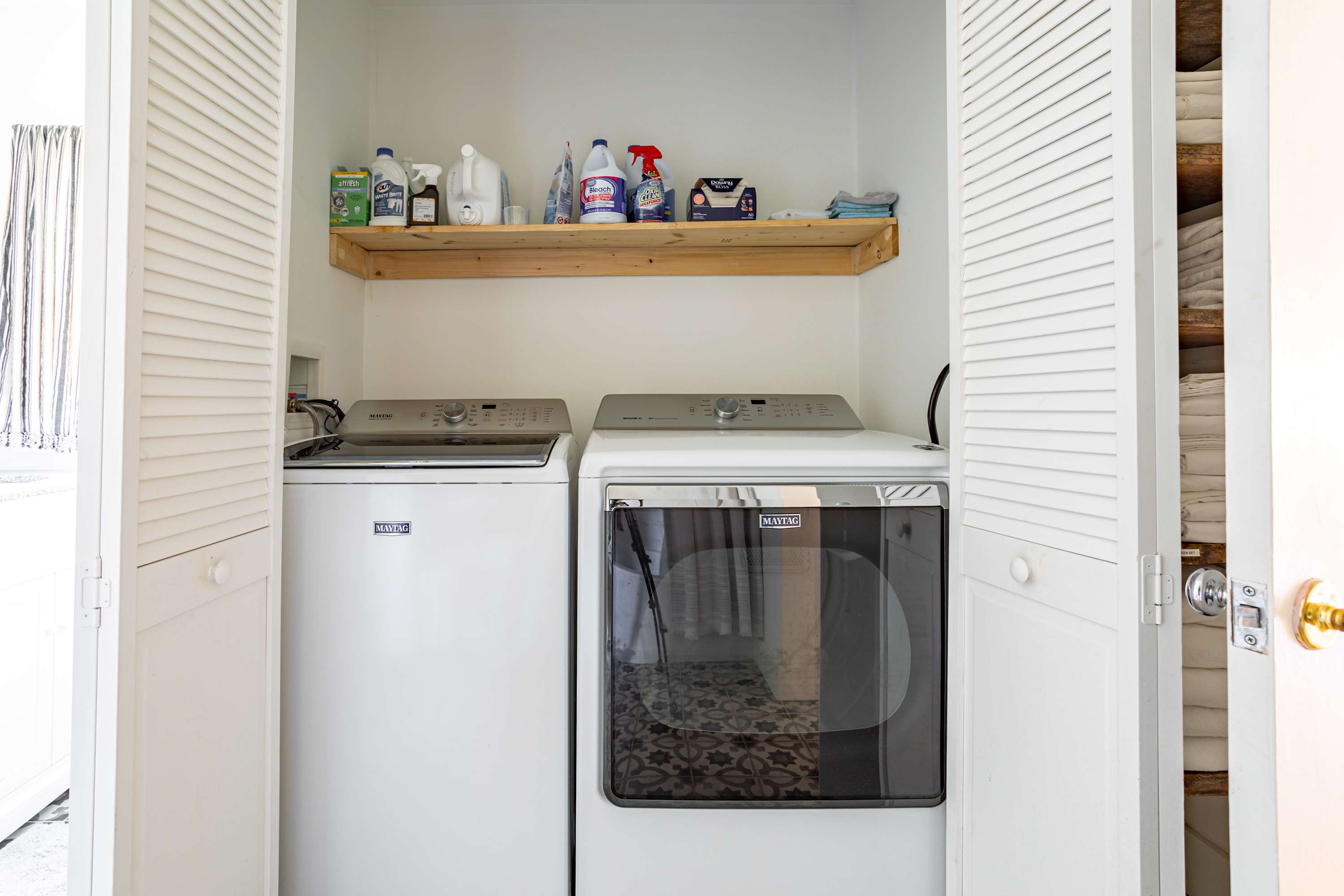 A compact laundry closet features a white top-load washing machine and a front-load dryer, neatly tucked behind open bi-fold doors. A rustic wooden shelf above holds various cleaning supplies and folded towels.