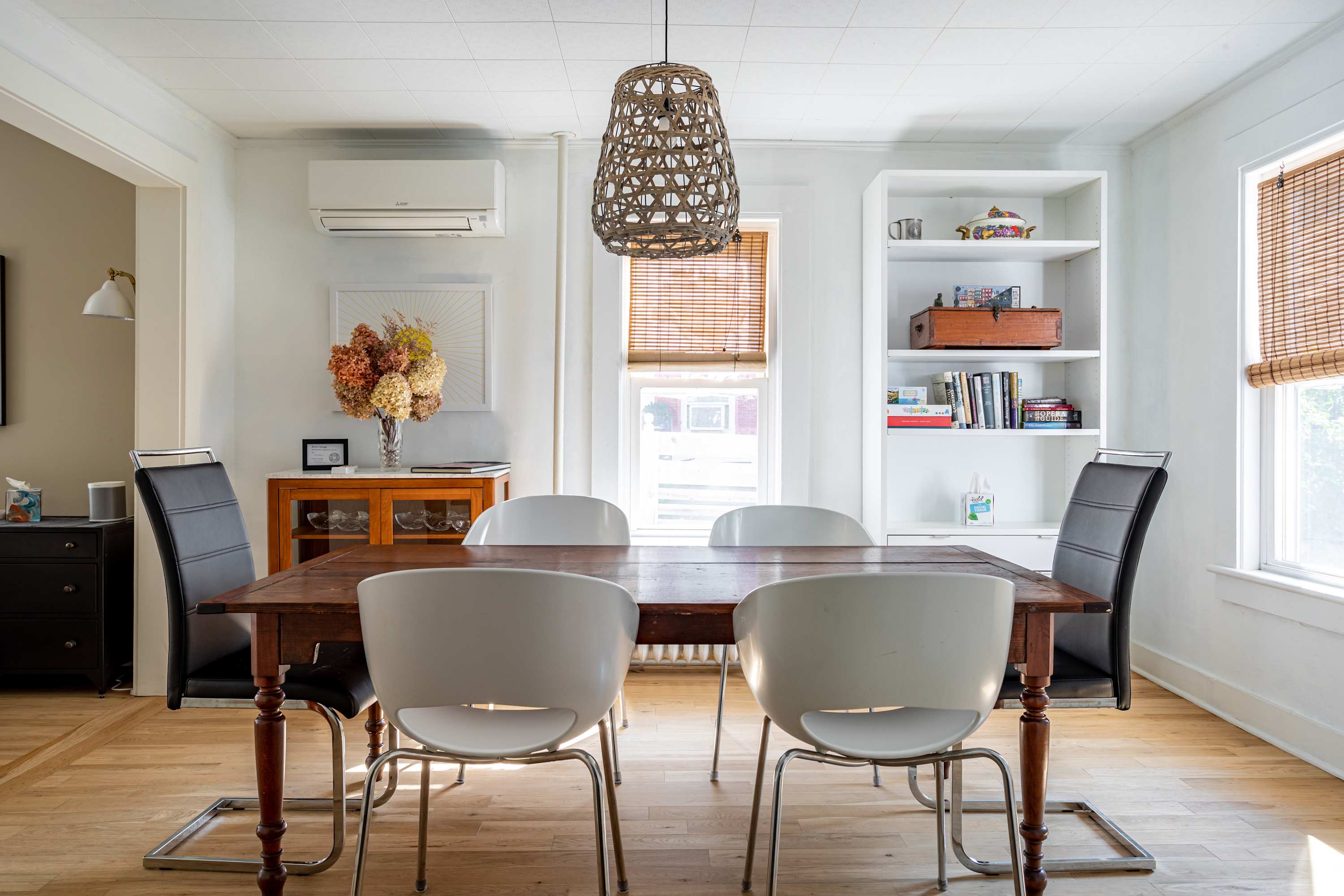 A bright dining room with a rustic wooden dining table surrounded by a mix of white and black chairs, illuminated by a woven pendant light, with built-in shelving and bamboo blinds on the windows.