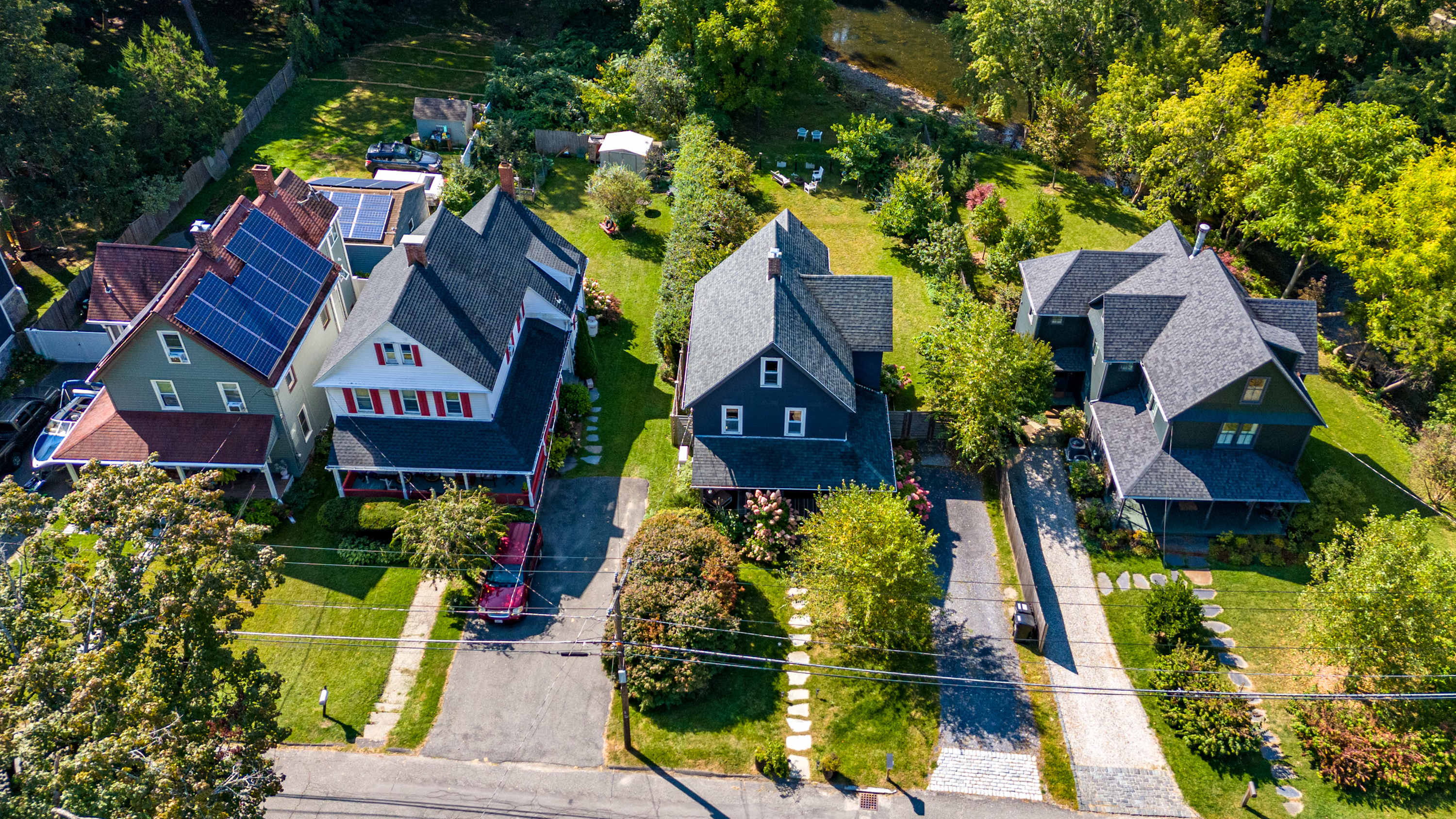 An aerial view showcases a residential street with several houses, including a dark gray property with a paved driveway and green lawn, surrounded by mature trees. A river is visible in the background, and solar panels are installed on a neighboring roof.