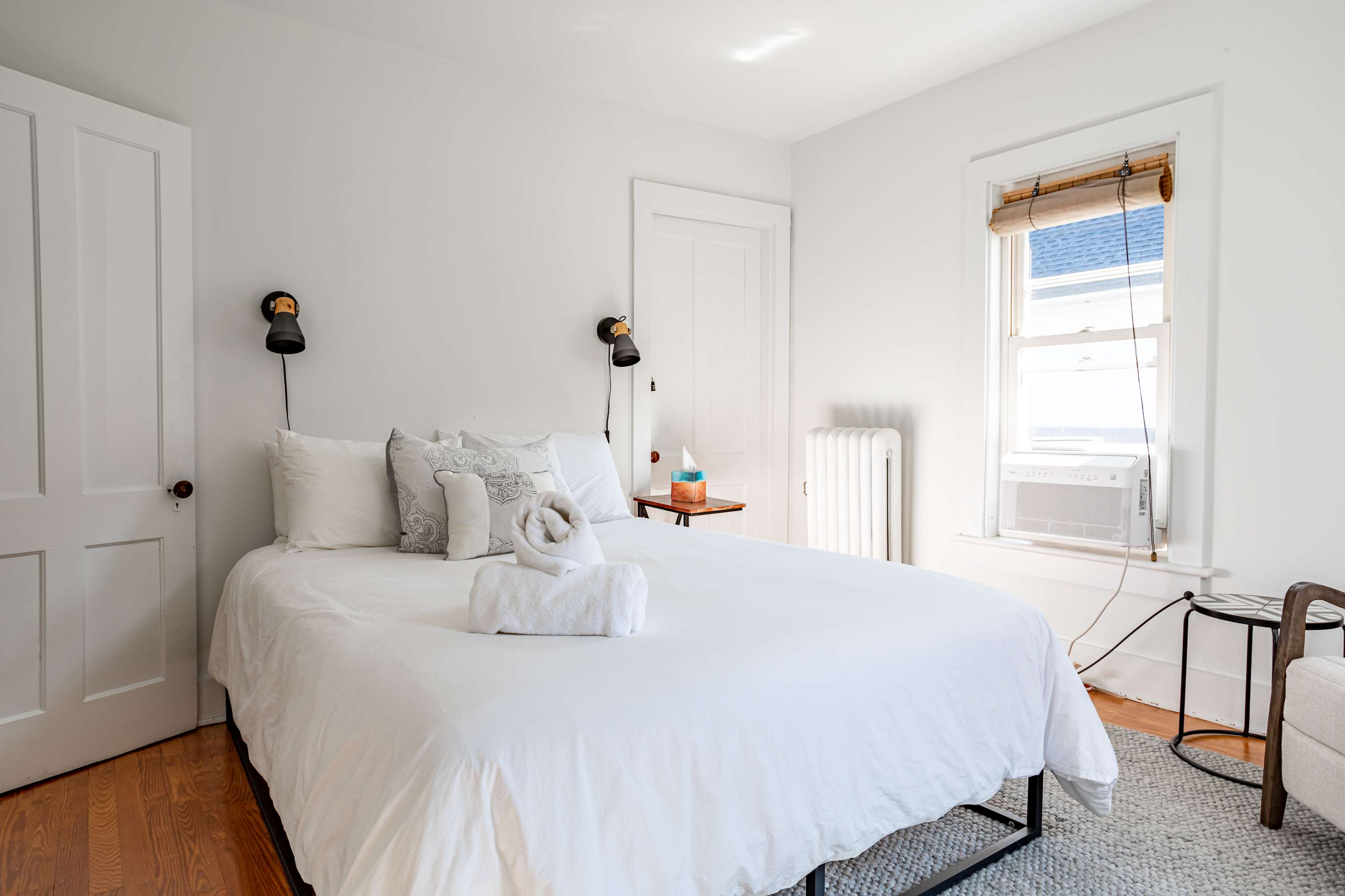 A bright bedroom features a queen-sized bed with crisp white bedding and two rolled towels, flanked by black wall-mounted sconces. A window with a window AC unit and a white radiator are visible on the right.