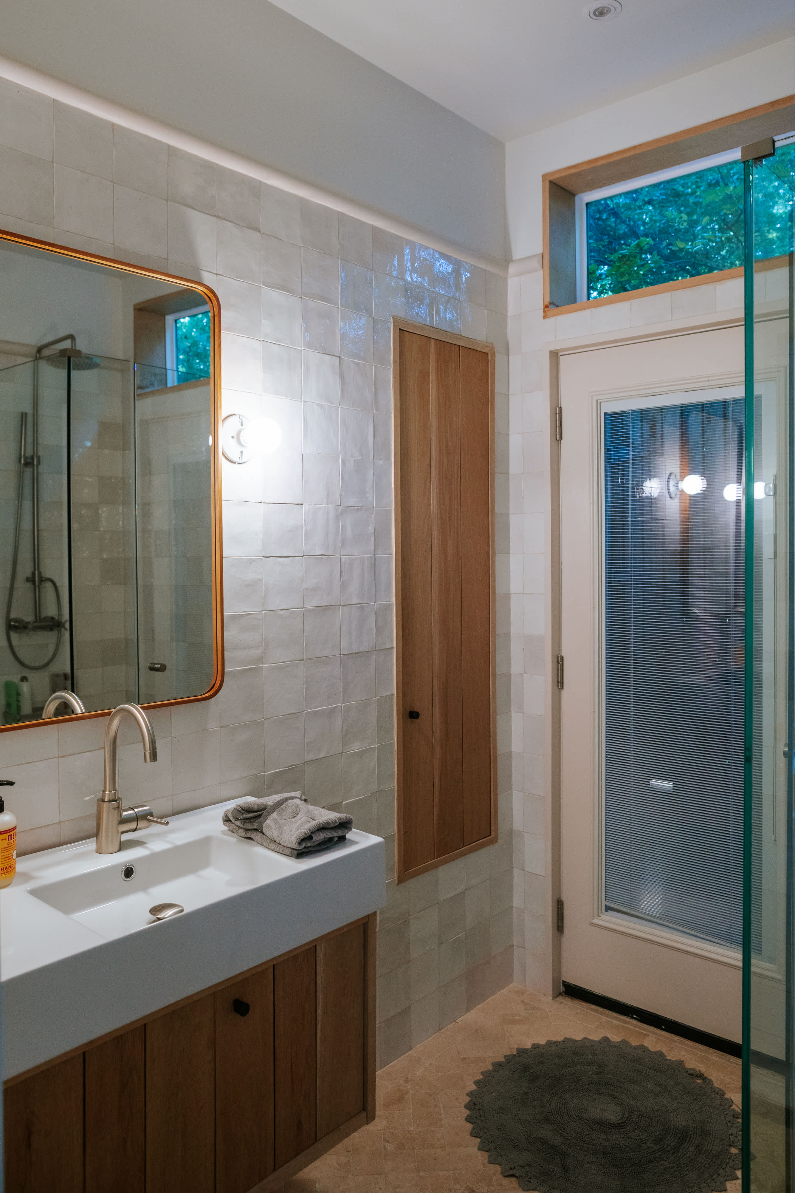 Contemporary bathroom with a white vanity, rectangular sink, and a gold-framed mirror. The walls are covered in light square tiles, and a glass shower enclosure is visible to the right.