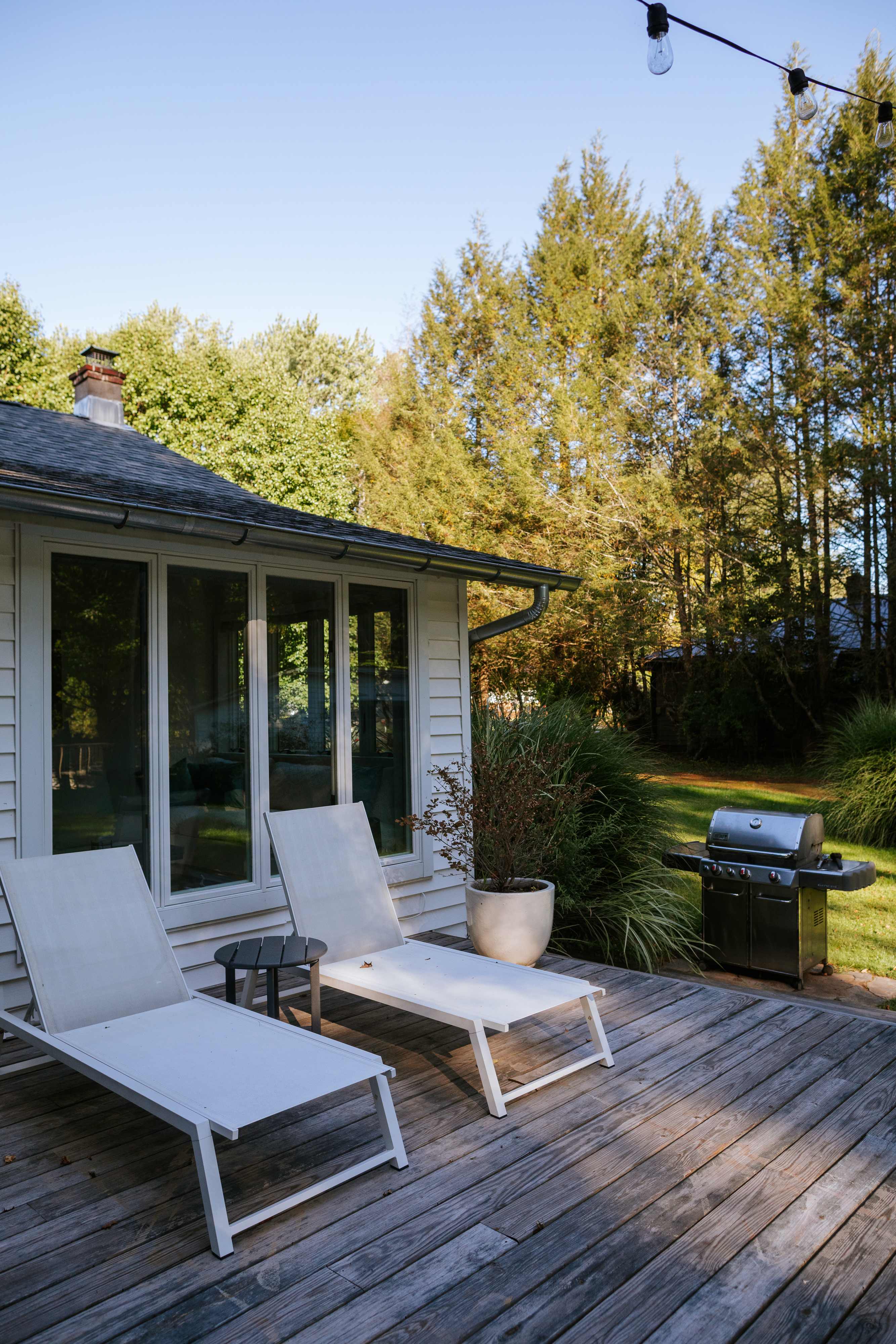 Outdoor deck with two white lounge chairs and a BBQ grill, set against a backdrop of tall evergreen trees. String lights are visible overhead, suggesting evening ambiance.