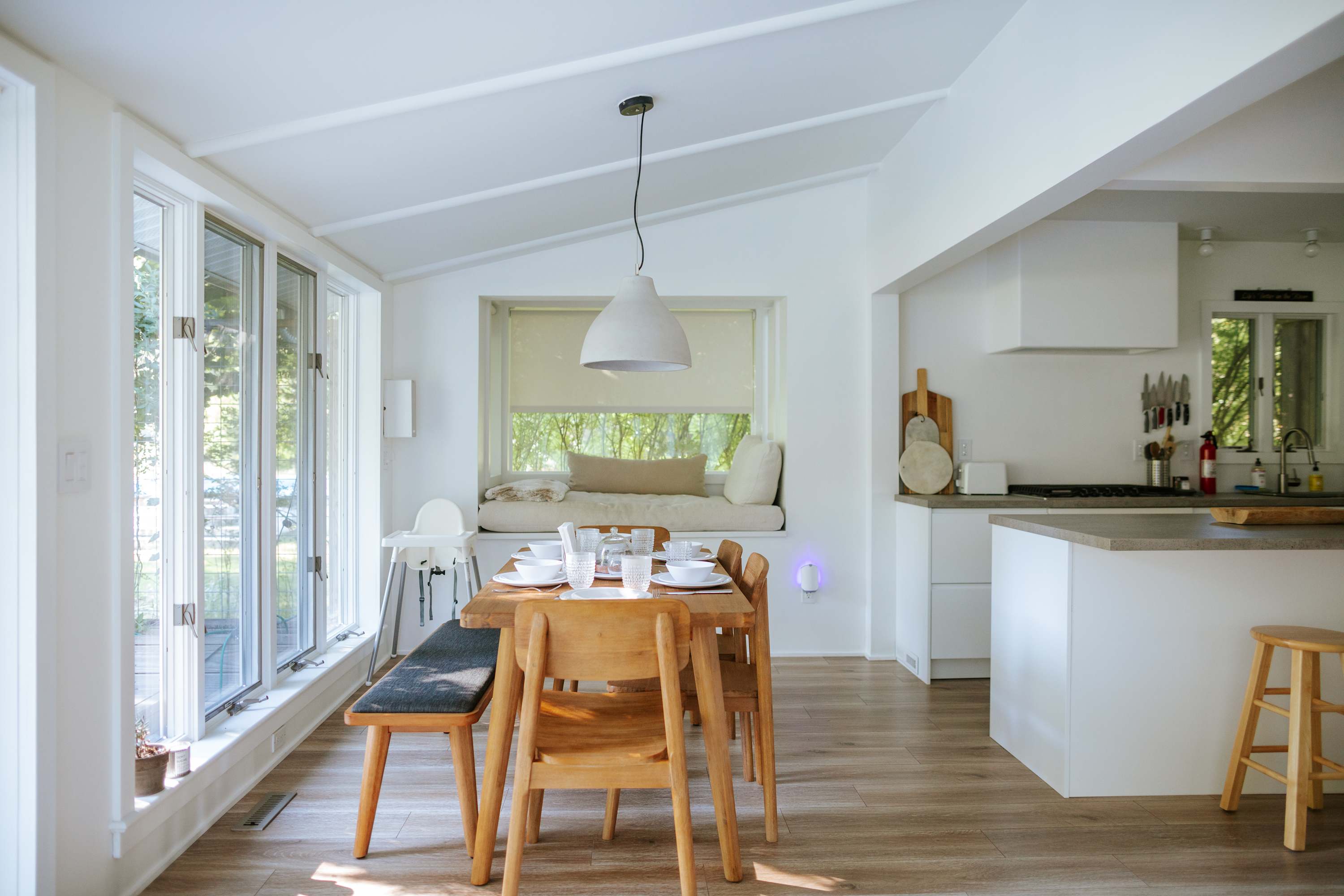 Bright dining area with a wooden table set for six, featuring a modern pendant light, large windows, and an adjacent built-in window seat with cushions, connecting to the kitchen.