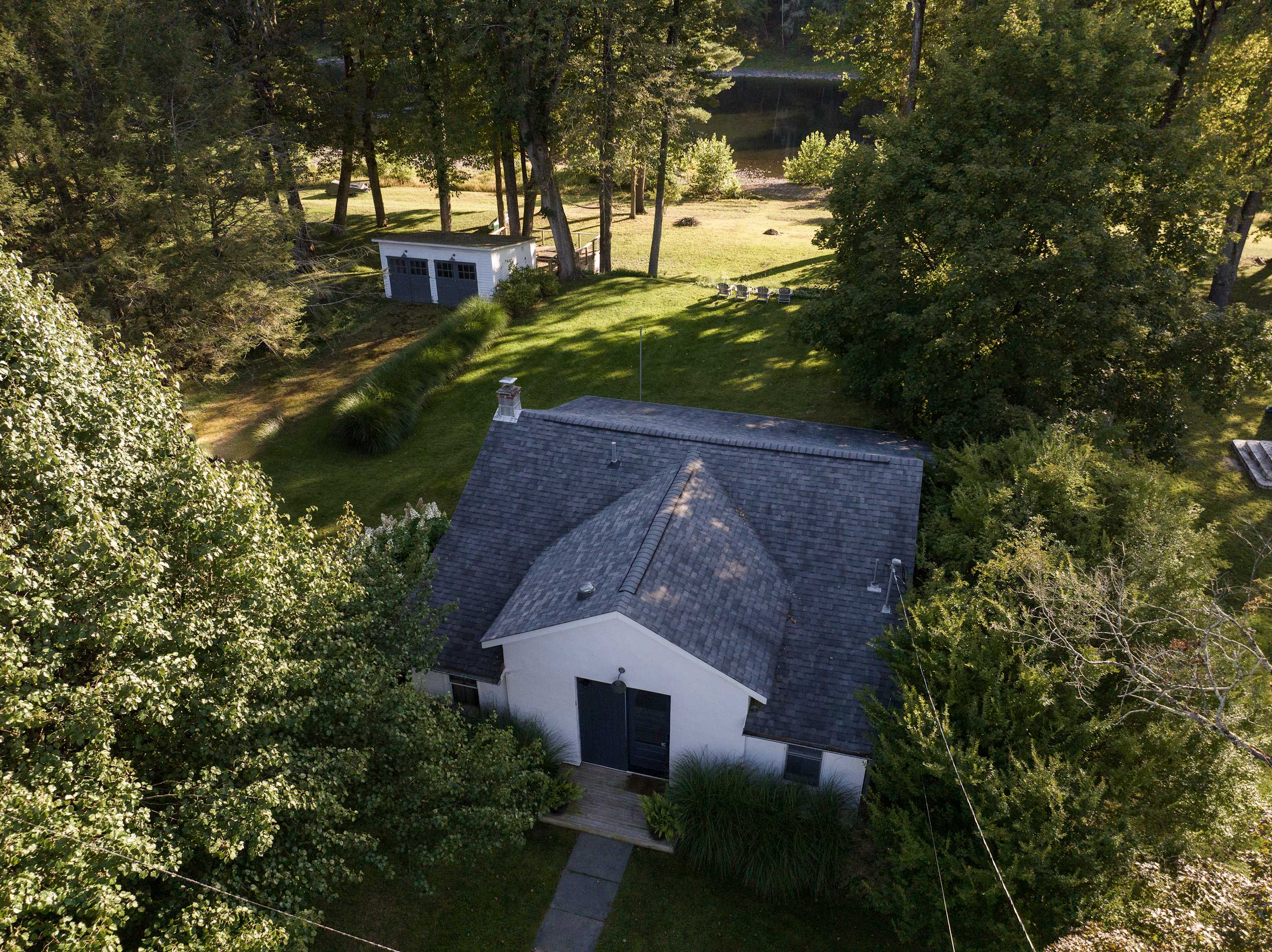 Aerial view of a white house with a dark gray roof, surrounded by lush green trees and grass. A small shed is visible in the background, with a glimpse of a body of water beyond the trees.