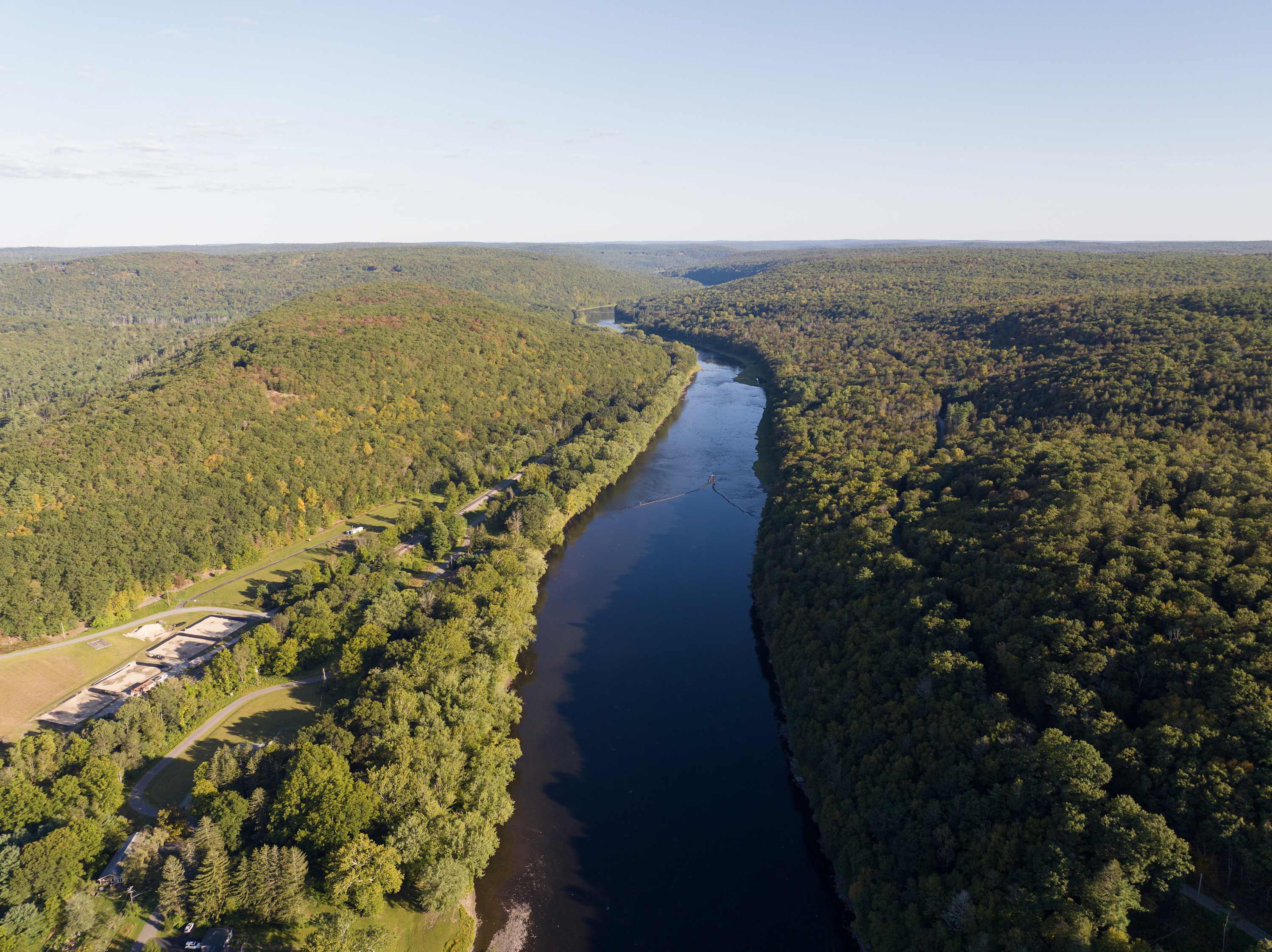 Expansive aerial view of a wide, dark river winding through a valley covered in dense green forests under a clear blue sky. The river appears calm, reflecting the sky.