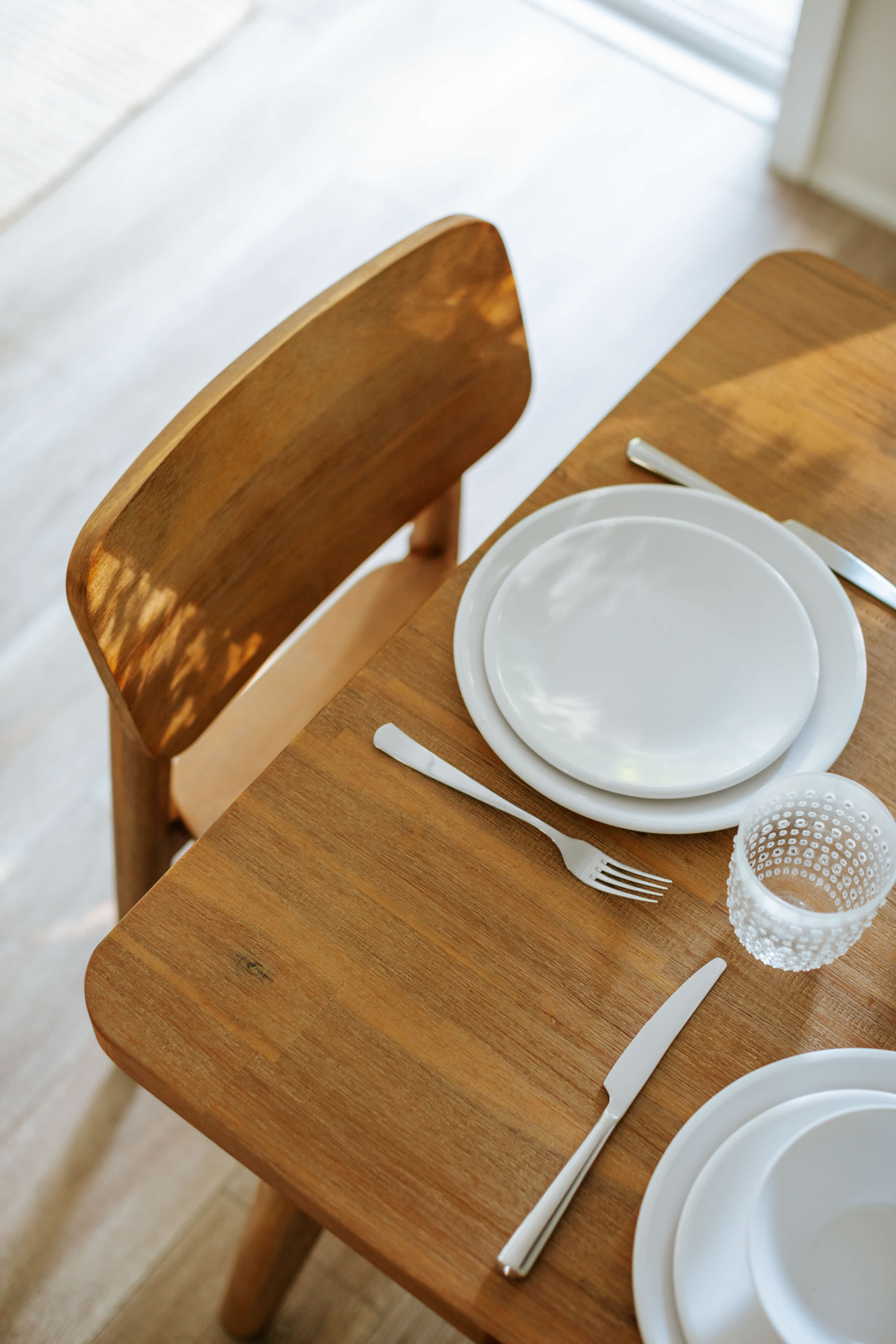 Detailed close-up of a single place setting on a light wooden dining table, featuring stacked white plates, a fork, knife, and a textured clear glass, with sunlight dappling the surface.