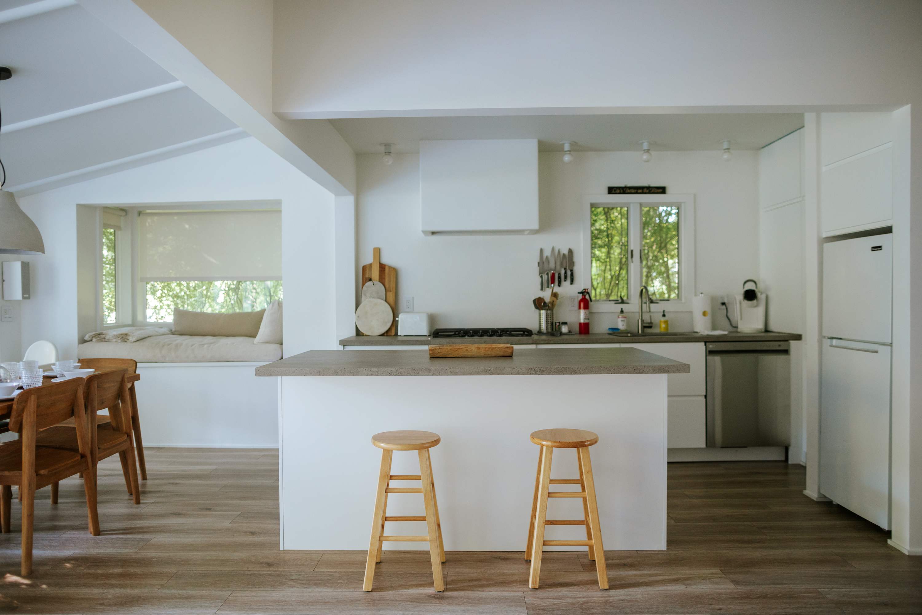 Modern kitchen with a light gray countertop island and two wooden bar stools, featuring white cabinetry, stainless steel appliances, and an open view to the adjacent dining area.