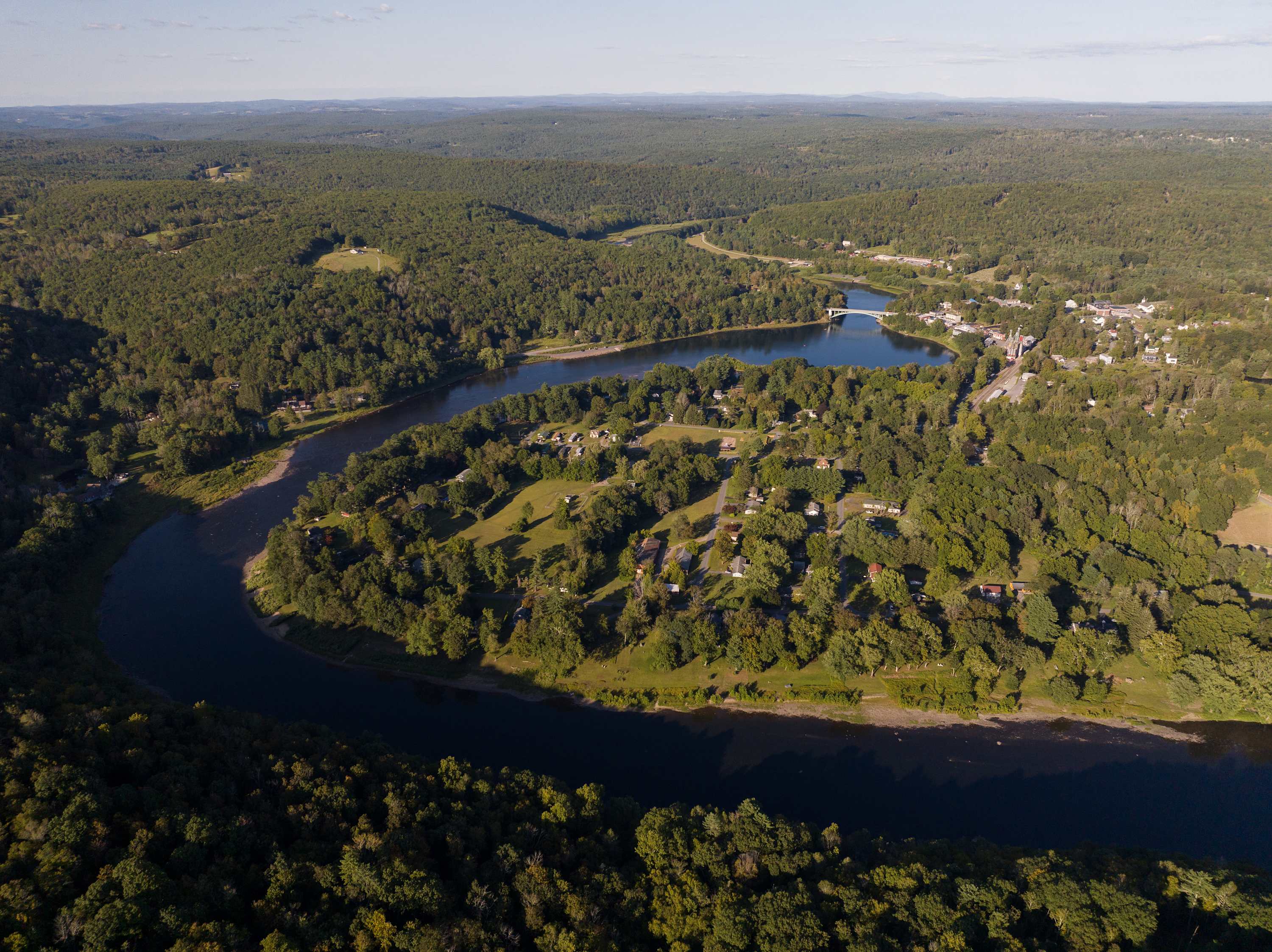 An expansive aerial view showcasing a winding river through a dense forest, with a small town and bridge visible in the distance, highlighting the property's secluded, natural setting.
