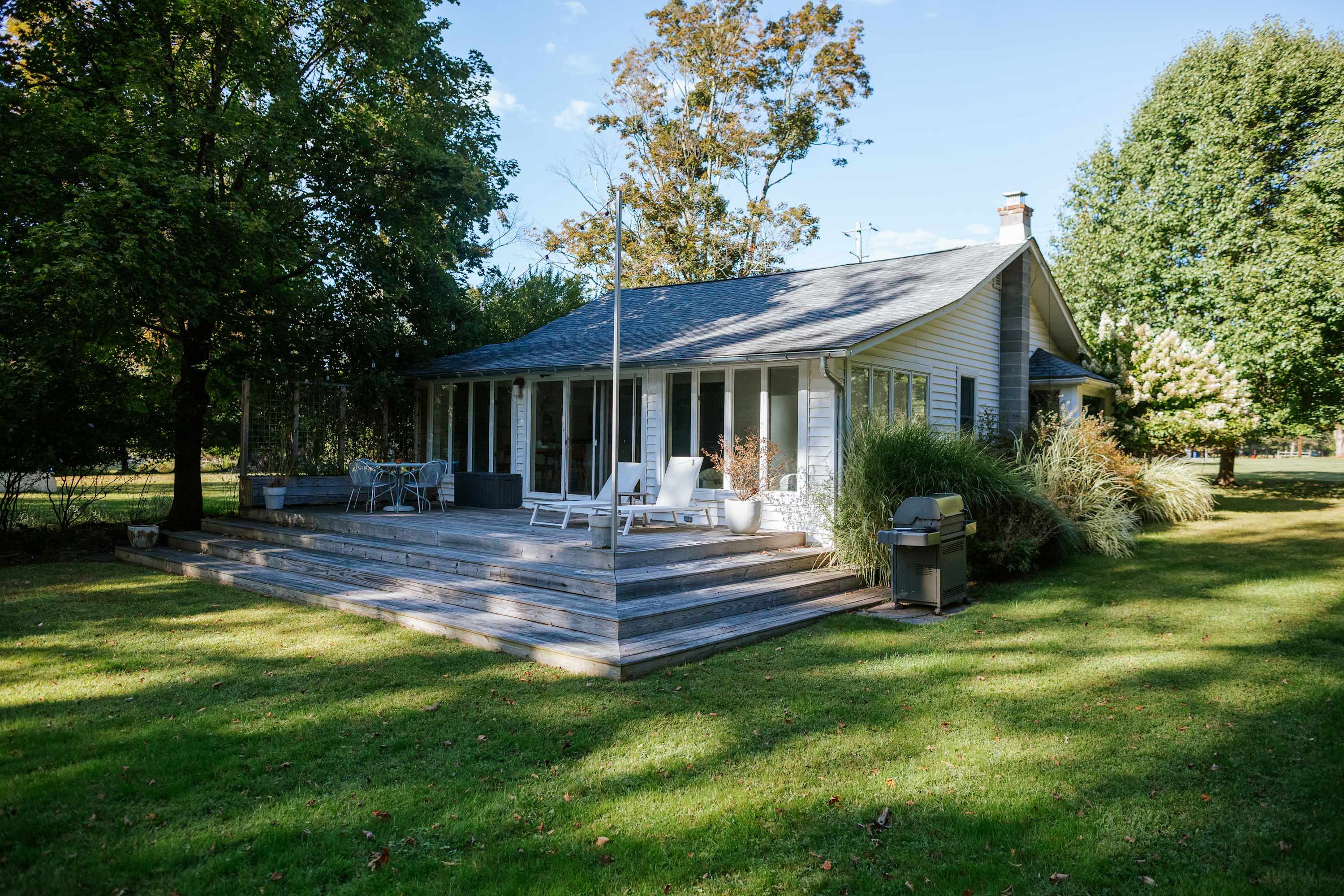 The back of a white single-story house with a gray shingled roof, featuring a large multi-level wooden deck furnished with outdoor seating and a BBQ grill, surrounded by a lush green lawn and mature trees under a clear sky.