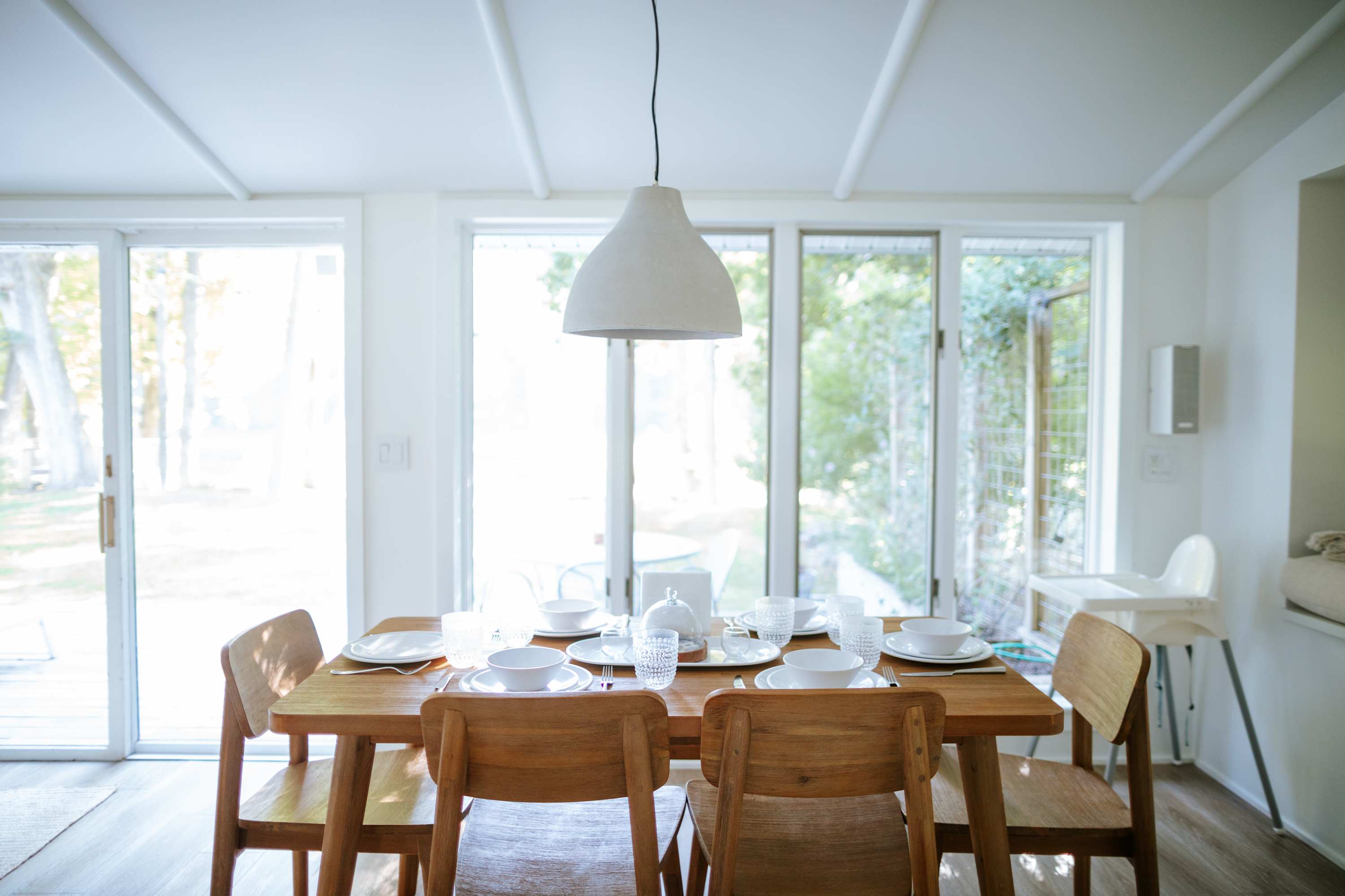 Close-up view of the wooden dining table, elegantly set with white plates, clear glasses, and silverware, illuminated by natural light from large windows.