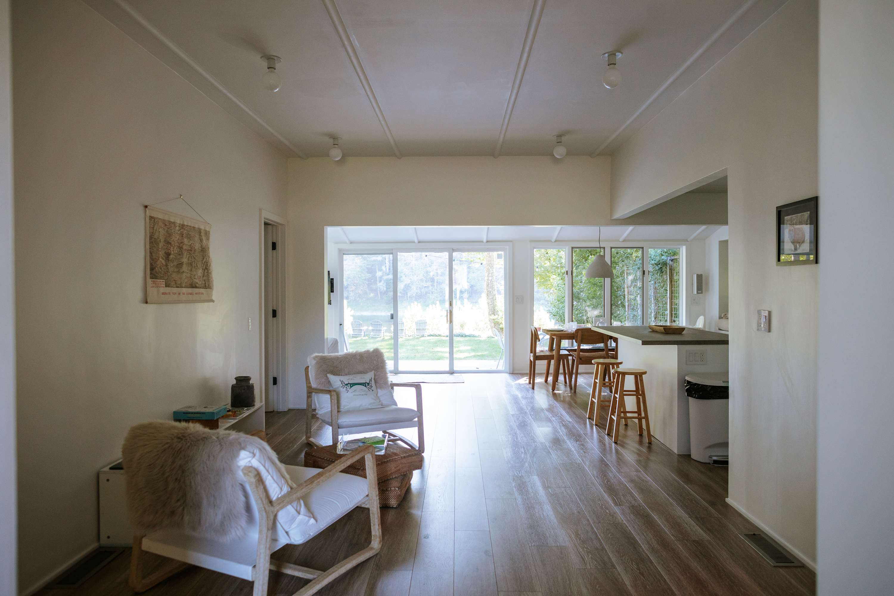 Bright, open-concept living space with light wood floors, two white armchairs with furry throws, and large glass doors leading to an outdoor area. A dining table and kitchen counter are visible in the background.