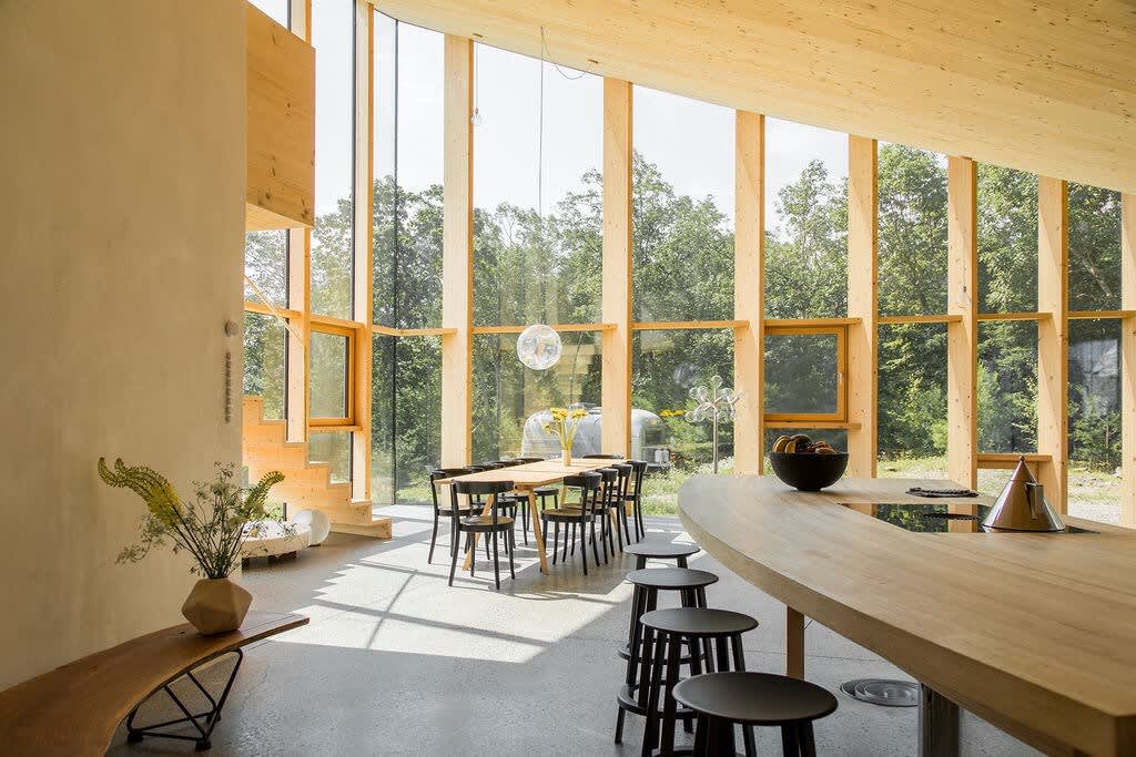 Modern dining area with a long wooden table and black chairs, set against a wall of floor-to-ceiling windows offering expansive views of lush trees. A curved wooden counter with bar stools and an integrated cooktop is in the foreground.