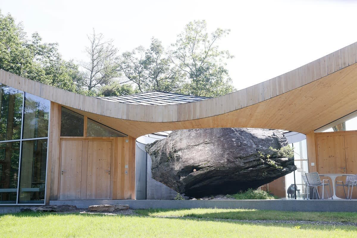 The house entrance features double wooden doors and a striking curved wooden canopy, leading to an open-air courtyard where a massive natural rock is dramatically integrated into the design, with outdoor seating nearby.