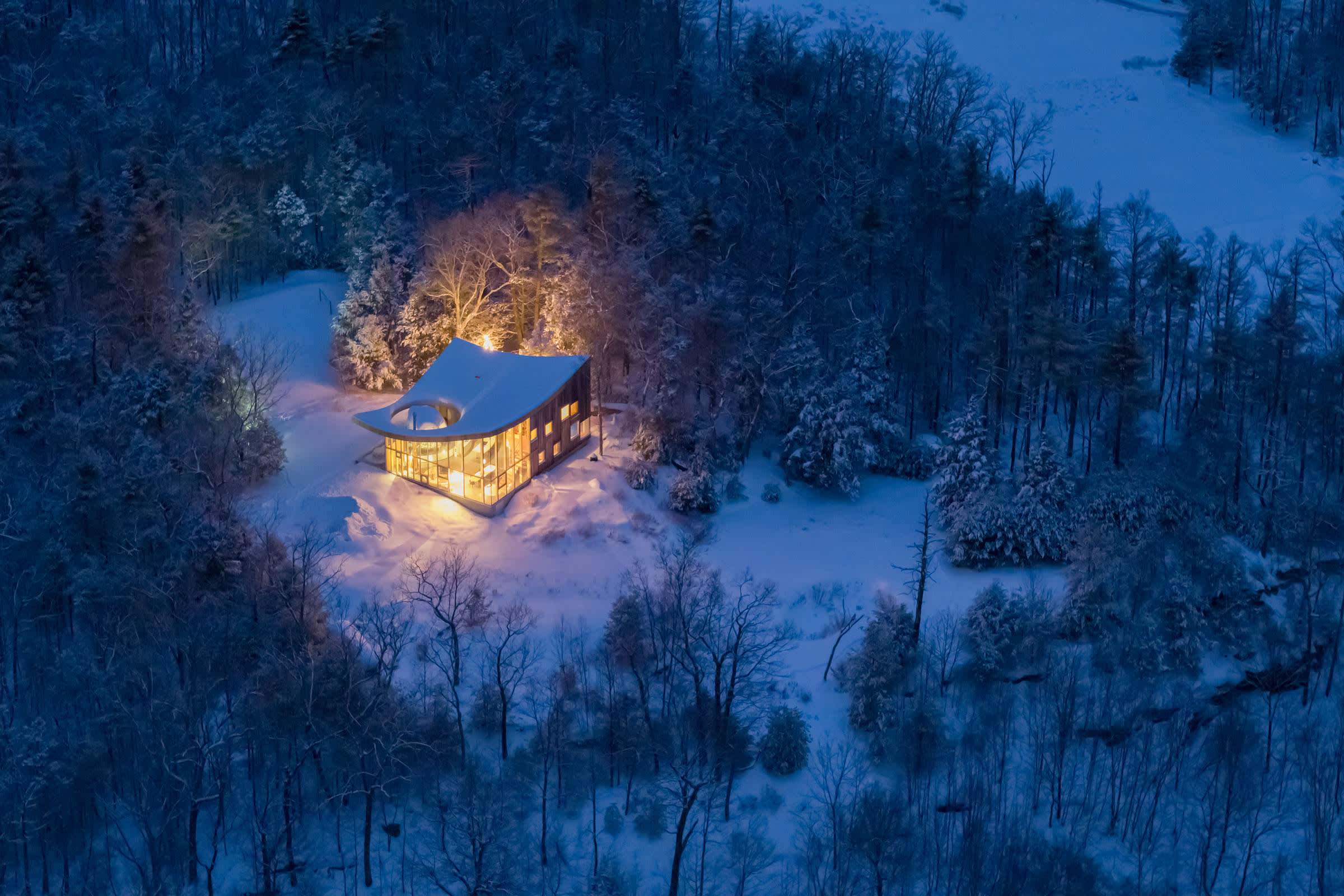 An enchanting aerial night view captures the warmly lit house nestled in a serene, snow-covered winter landscape, surrounded by bare and evergreen trees under a dark blue sky.