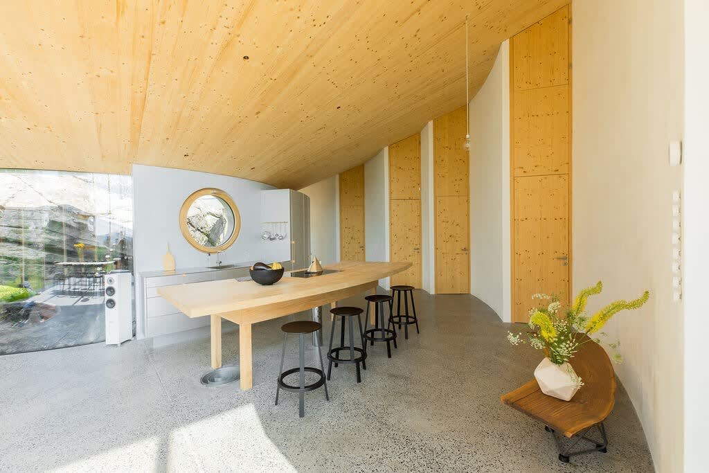 Expansive open-plan kitchen and dining area with a curved wooden ceiling and polished concrete floors. A long wooden island with bar stools is central, with a circular window in the background and a large glass wall revealing a rocky landscape.